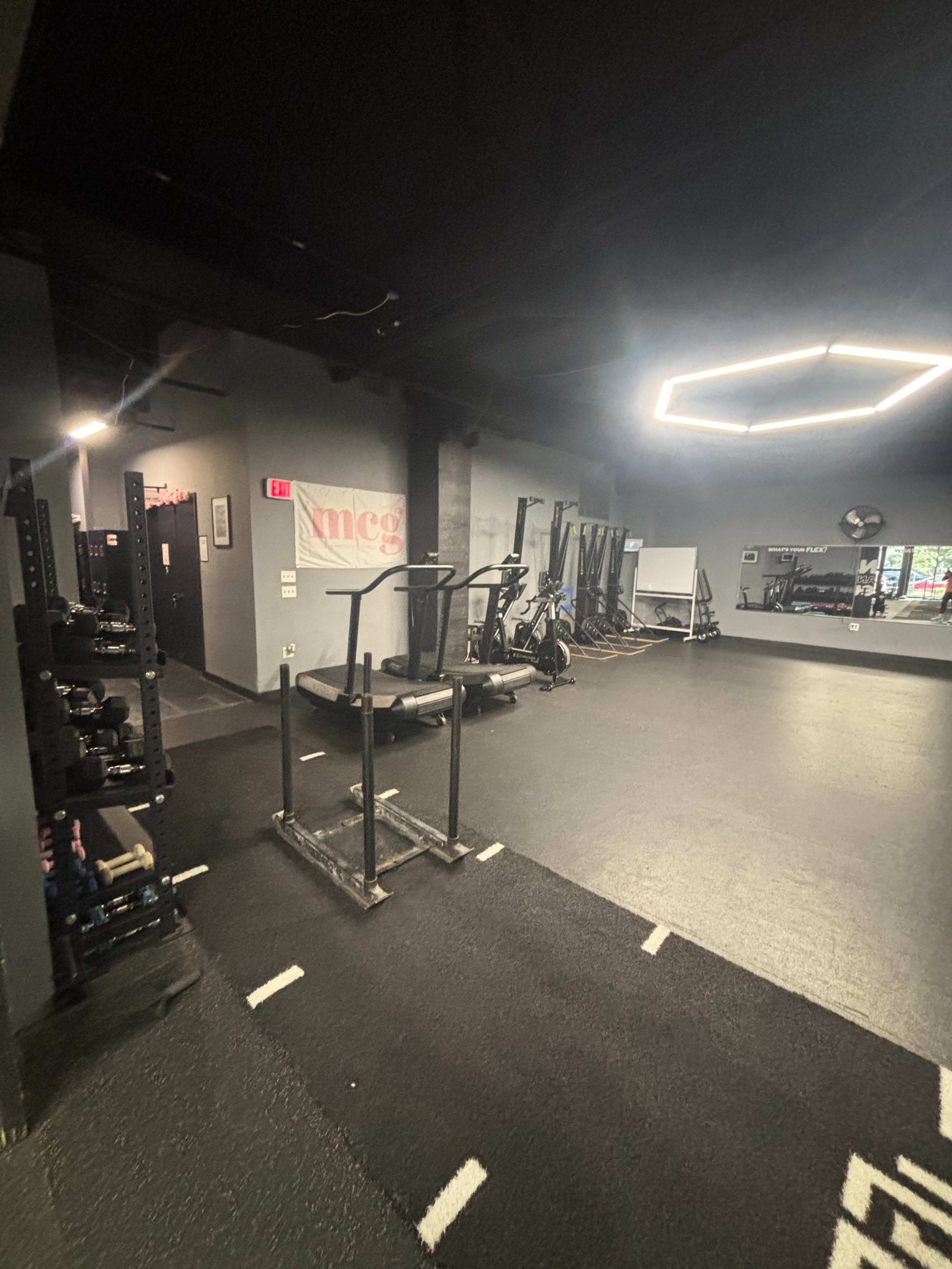 The image shows an empty gym space featuring fitness equipment, including a sled and exercise machines, with a mirrored wall and a banner in the background.