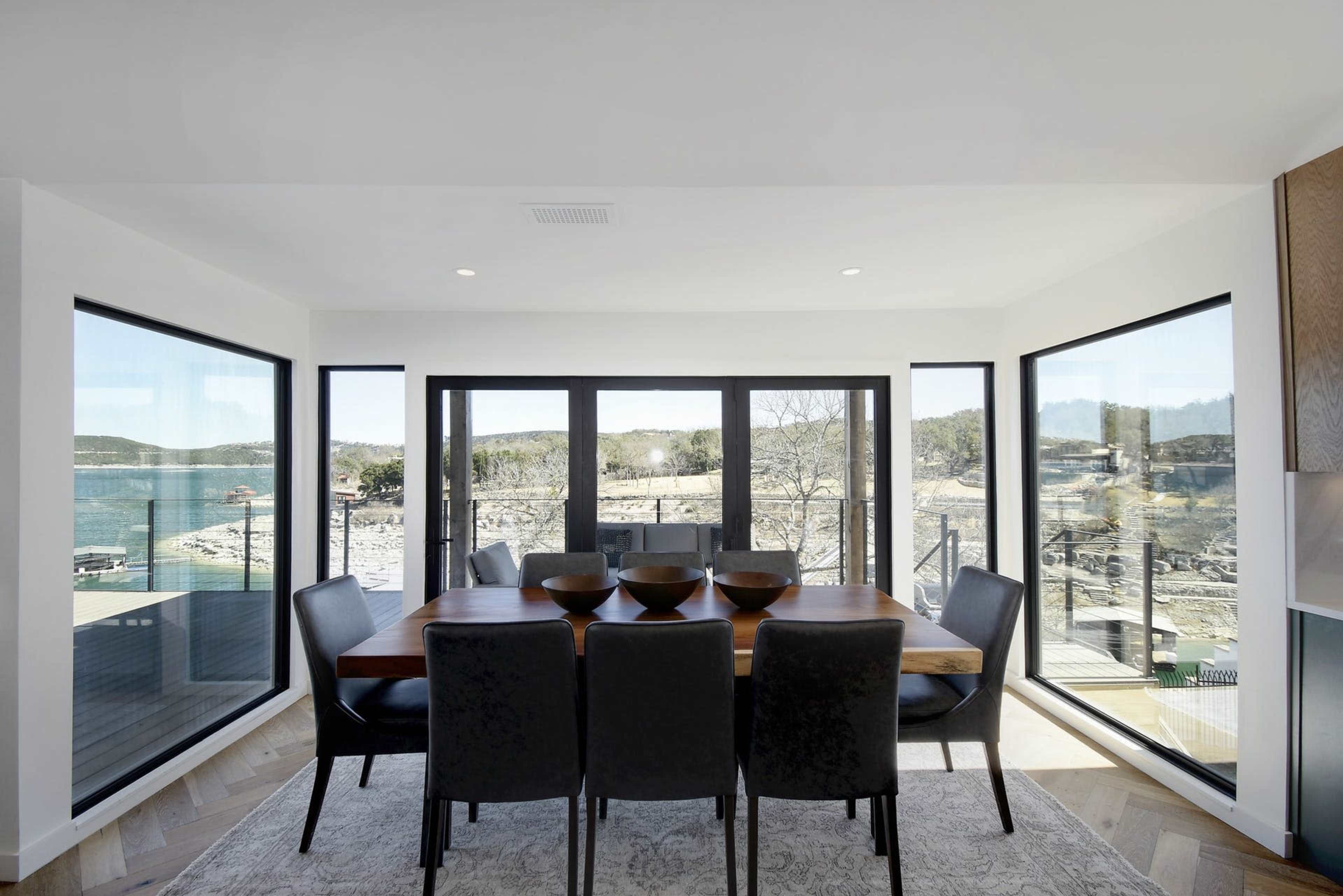 A modern dining area features a large table surrounded by black chairs, with expansive windows showcasing a view of a lake and rocky shoreline.