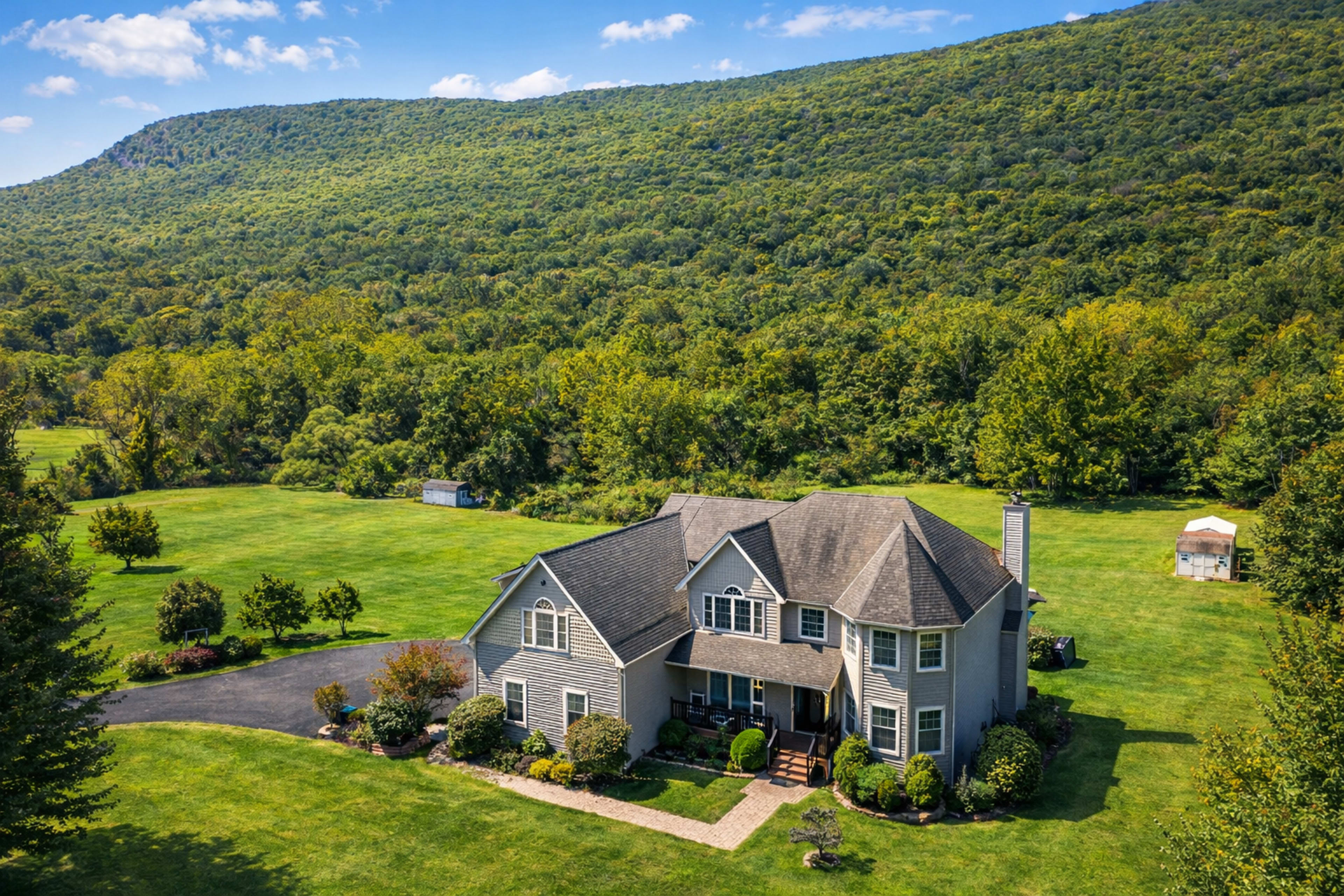 A large gray house is situated on a green, spacious lawn, surrounded by trees and set against a mountainous backdrop.