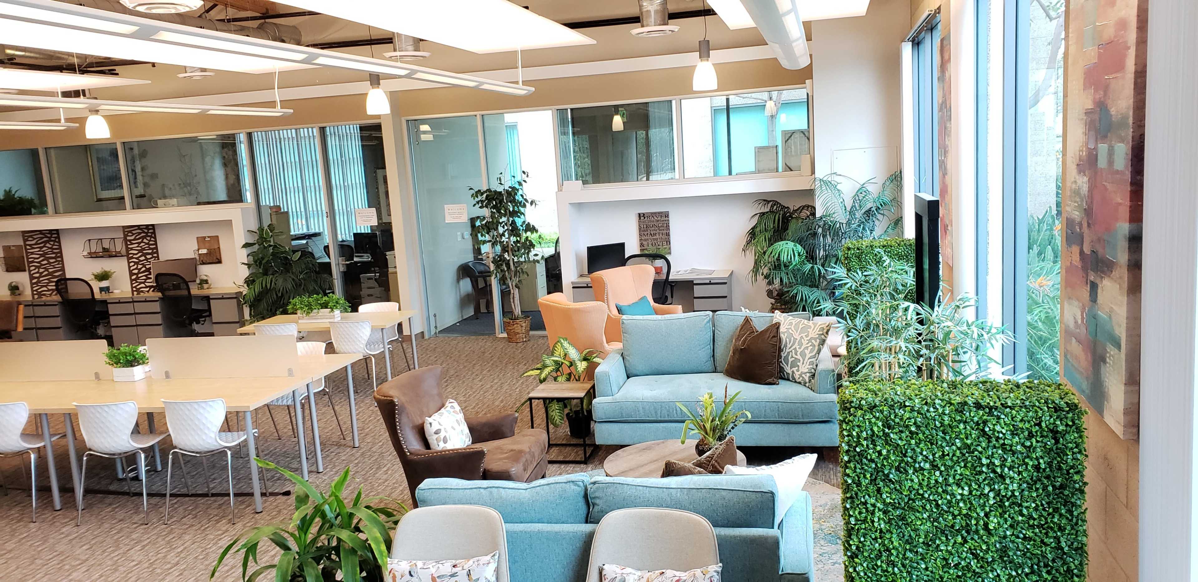A modern office space featuring a variety of seating arrangements, greenery, and workstations organized around a large communal table.