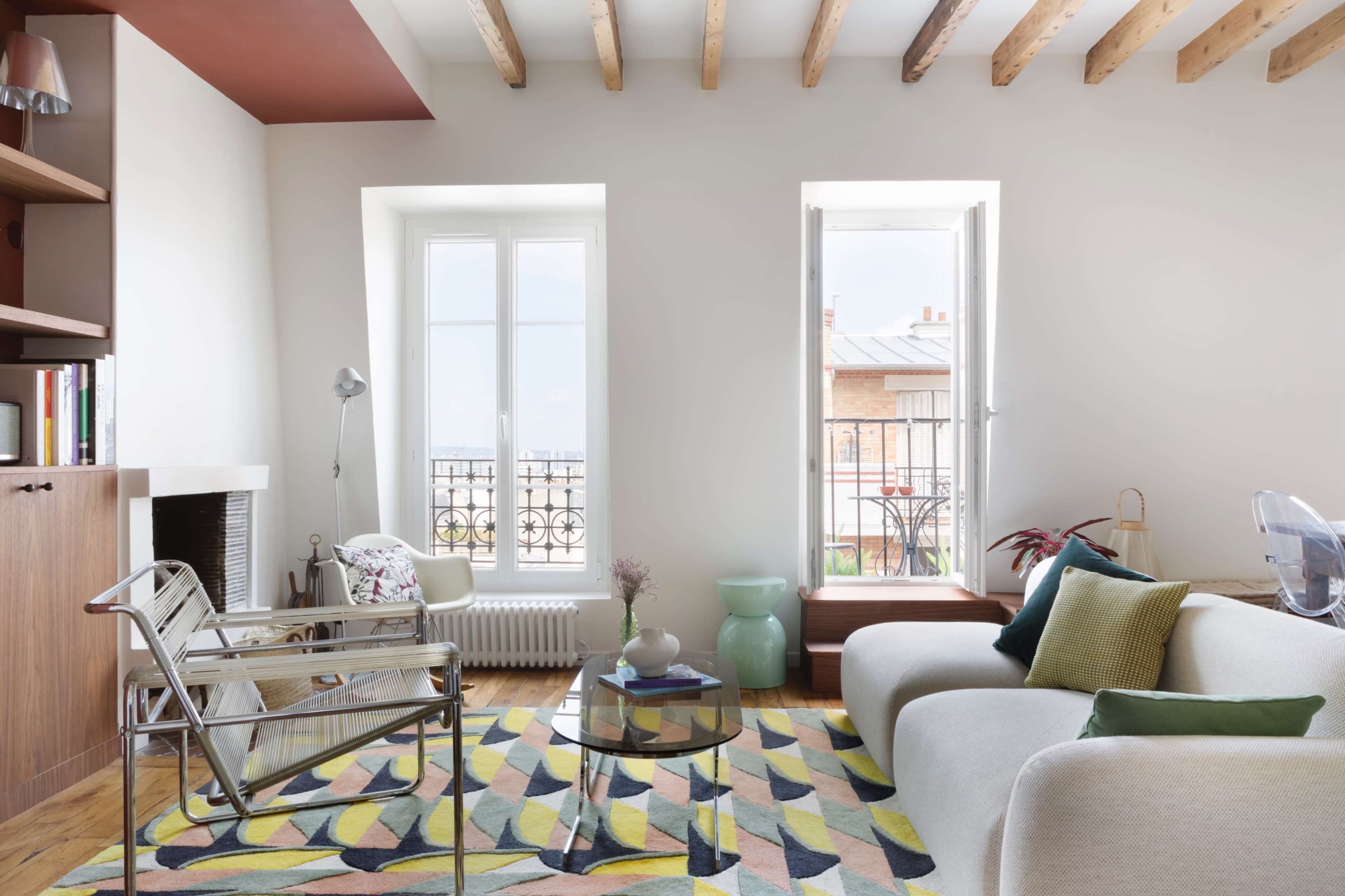 The image shows a modern living room with a light-colored sofa, a glass coffee table, and two large windows that overlook a balcony.