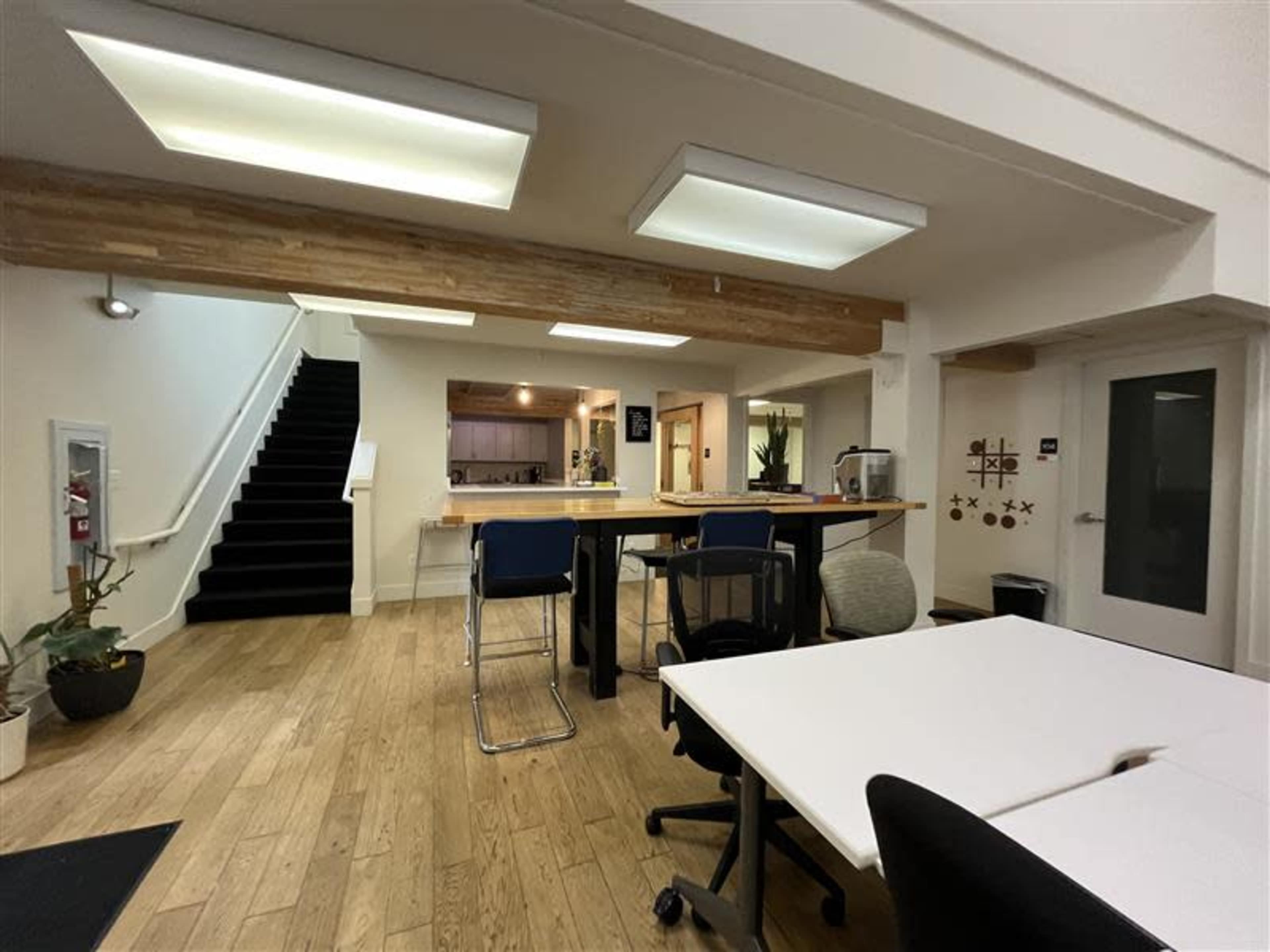 The image shows a modern office space with wooden flooring, a staircase in the background, and a communal table surrounded by chairs.