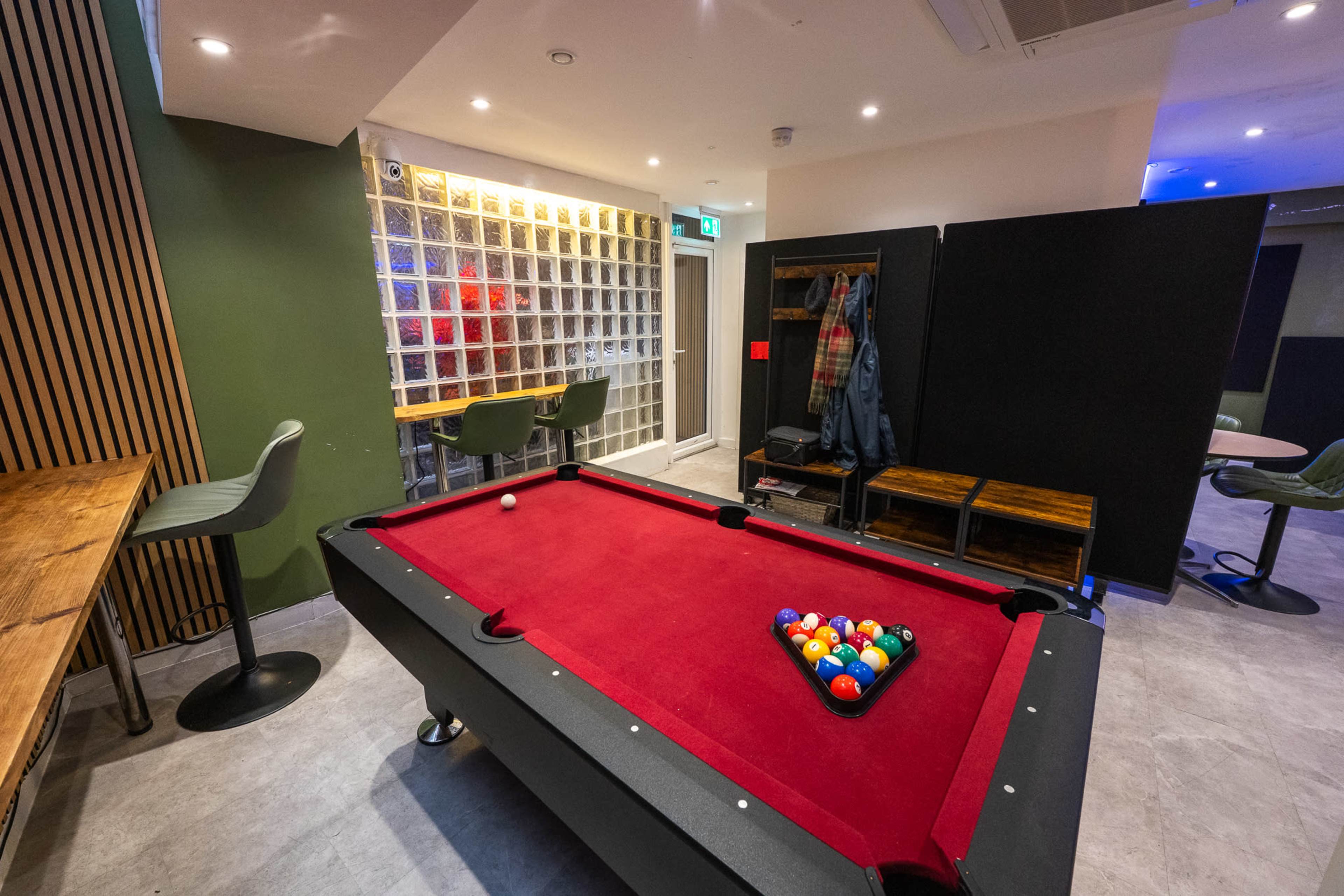 The image shows a modern recreational room featuring a red felt pool table, surrounded by seating areas and a coat rack in a well-lit space.