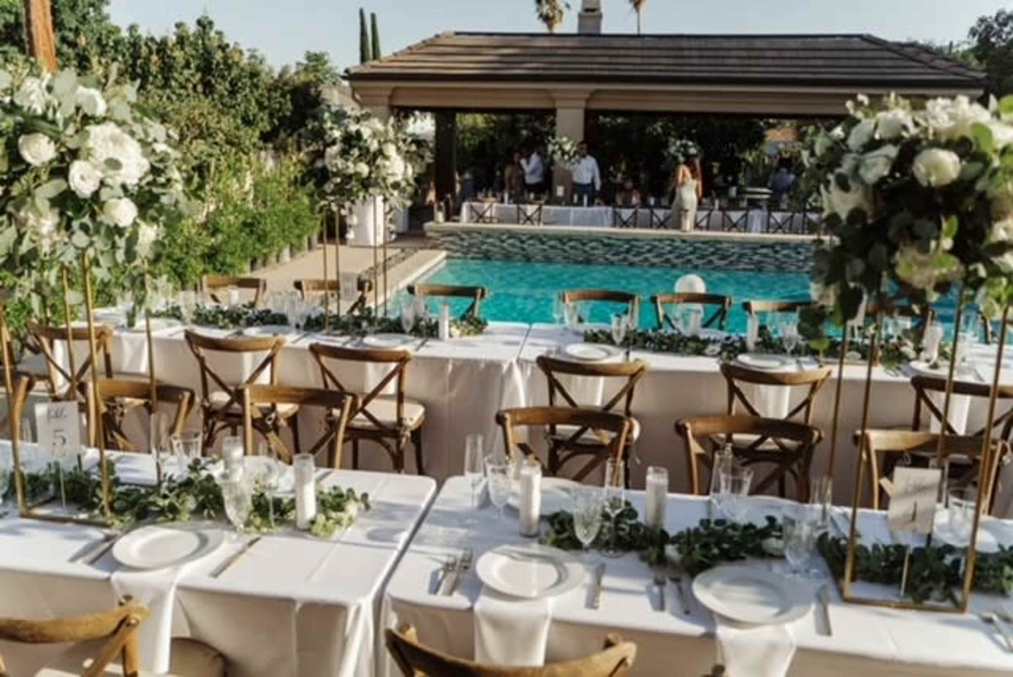 A long dining table is set outdoors by a swimming pool, adorned with floral arrangements and greenery.
