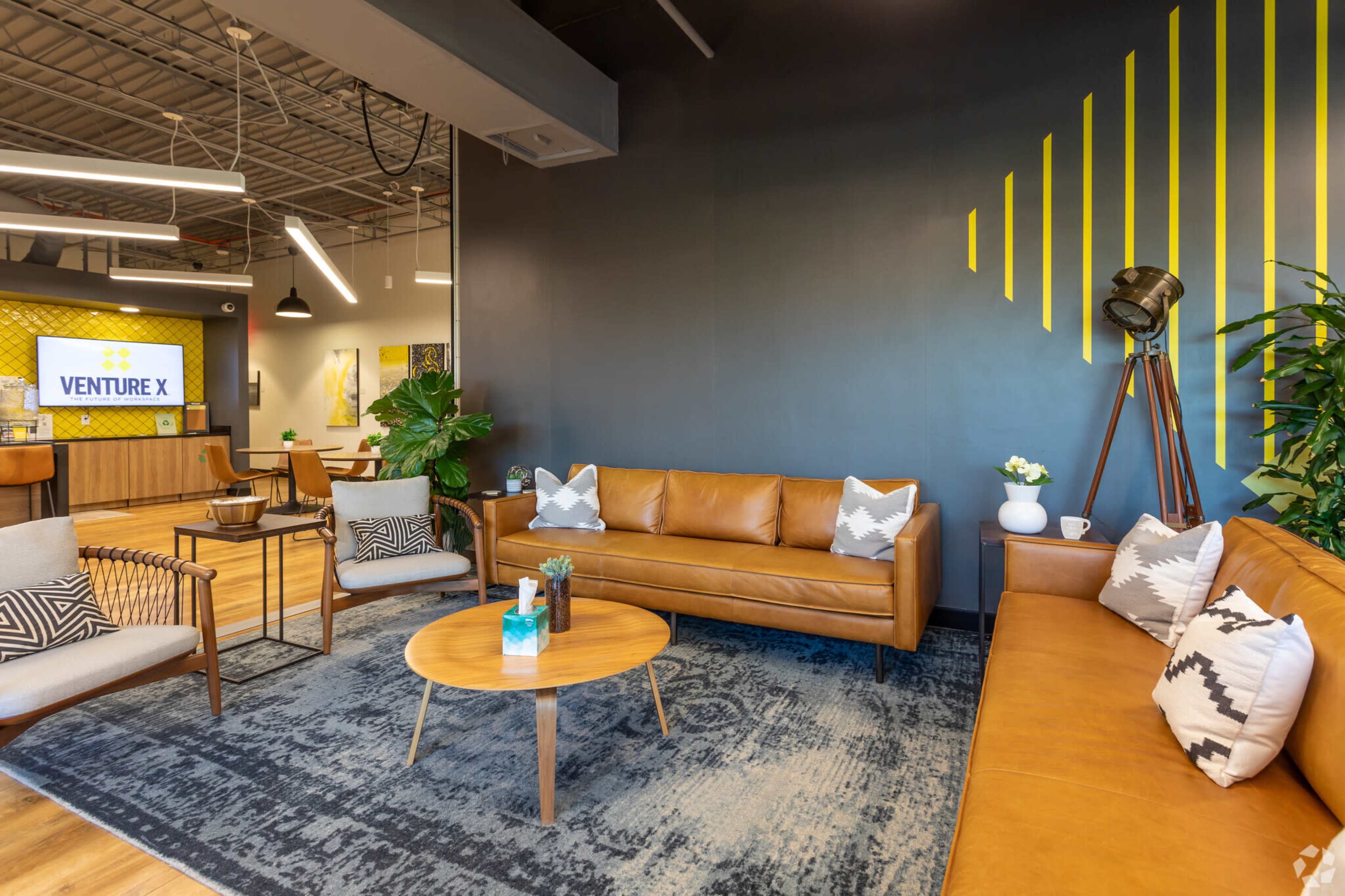 The image shows a modern office lounge area with brown leather sofas, a round wooden coffee table, and plants, set against a dark wall with yellow geometric decorations.