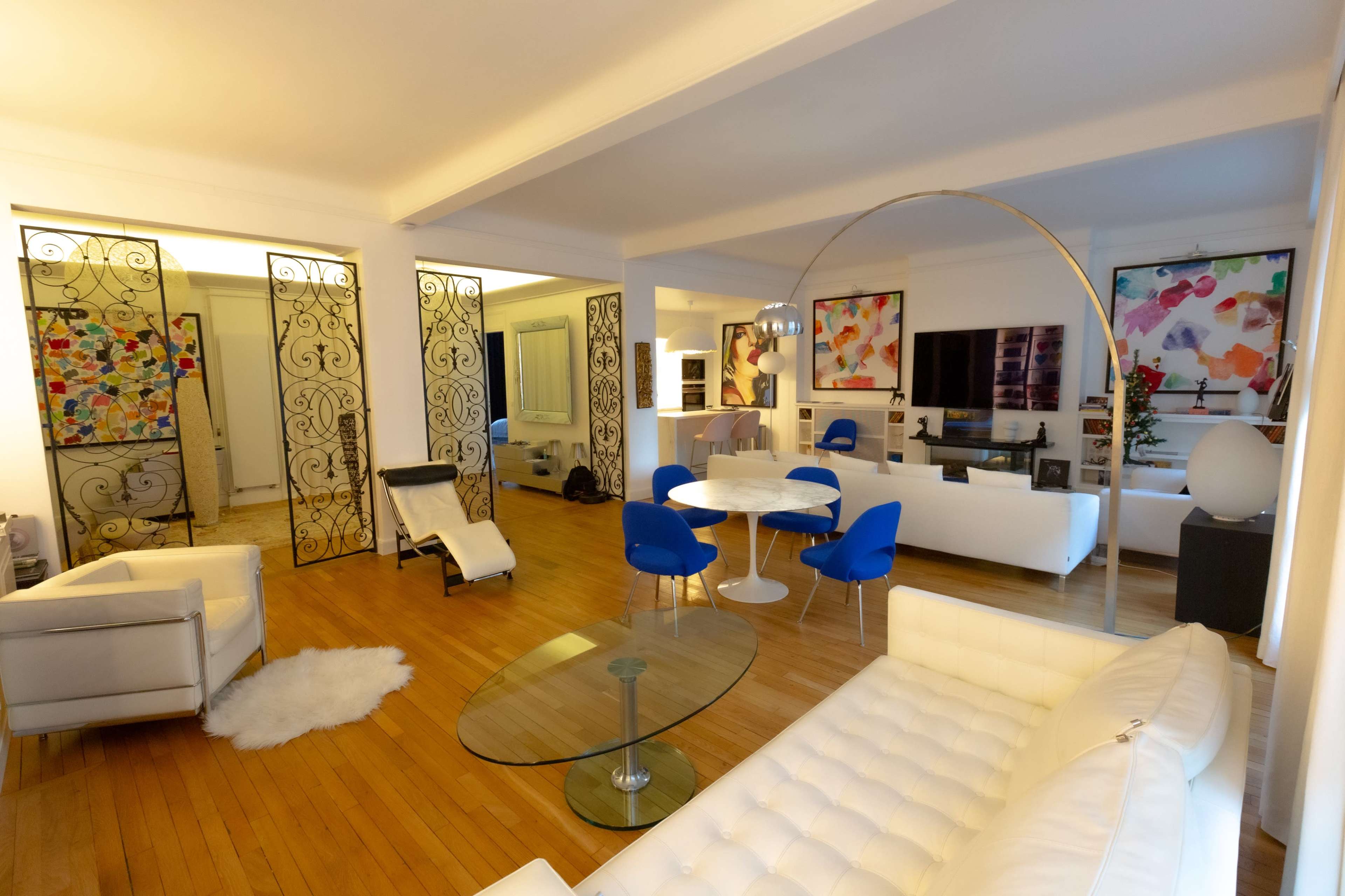 The image depicts a modern living room featuring a white leather sectional sofa, a circular dining table with blue chairs, and large abstract art pieces on the walls.