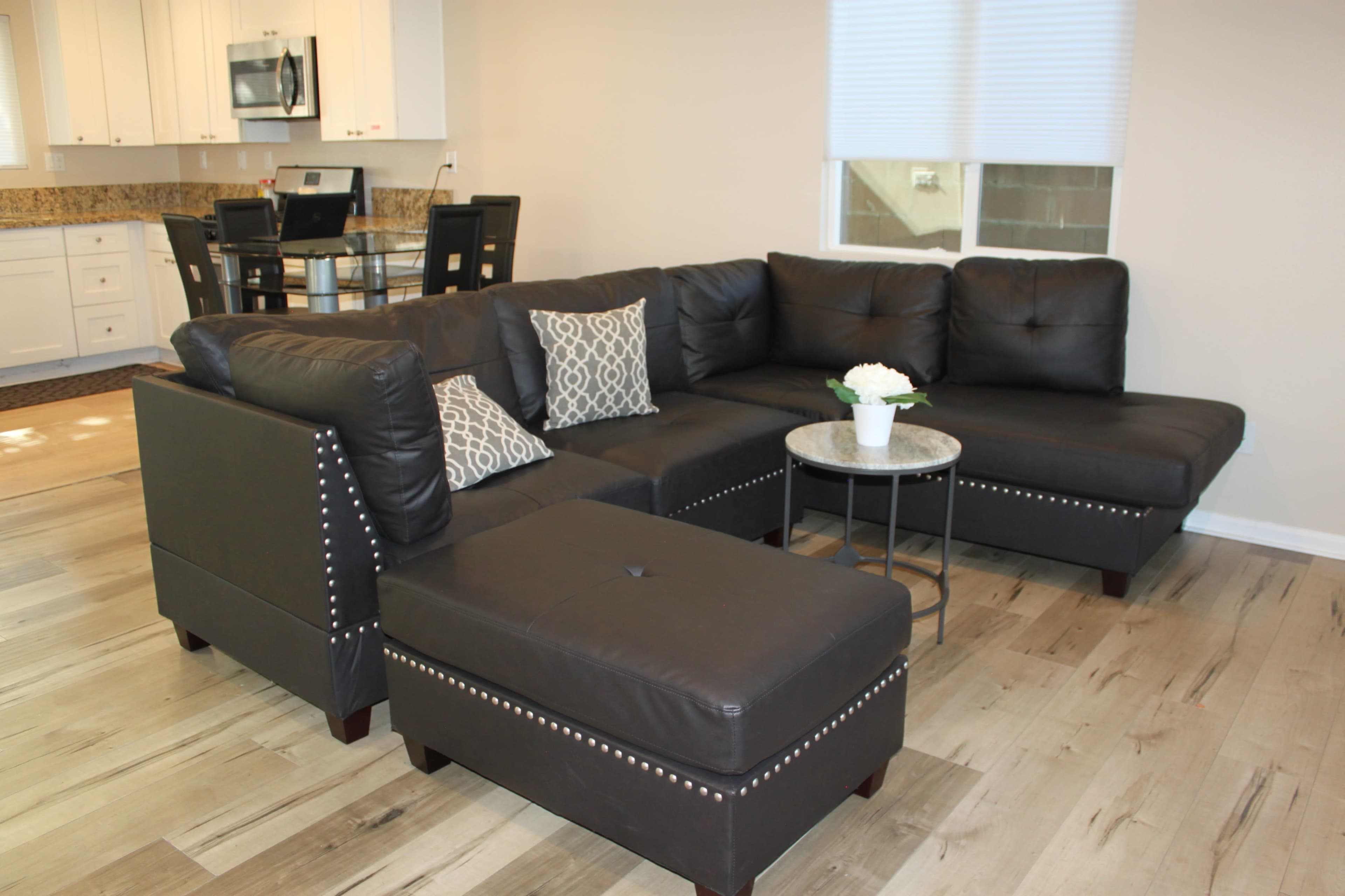 A modern living room featuring a dark sectional couch with decorative pillows, a round coffee table, and a dining area in the background.