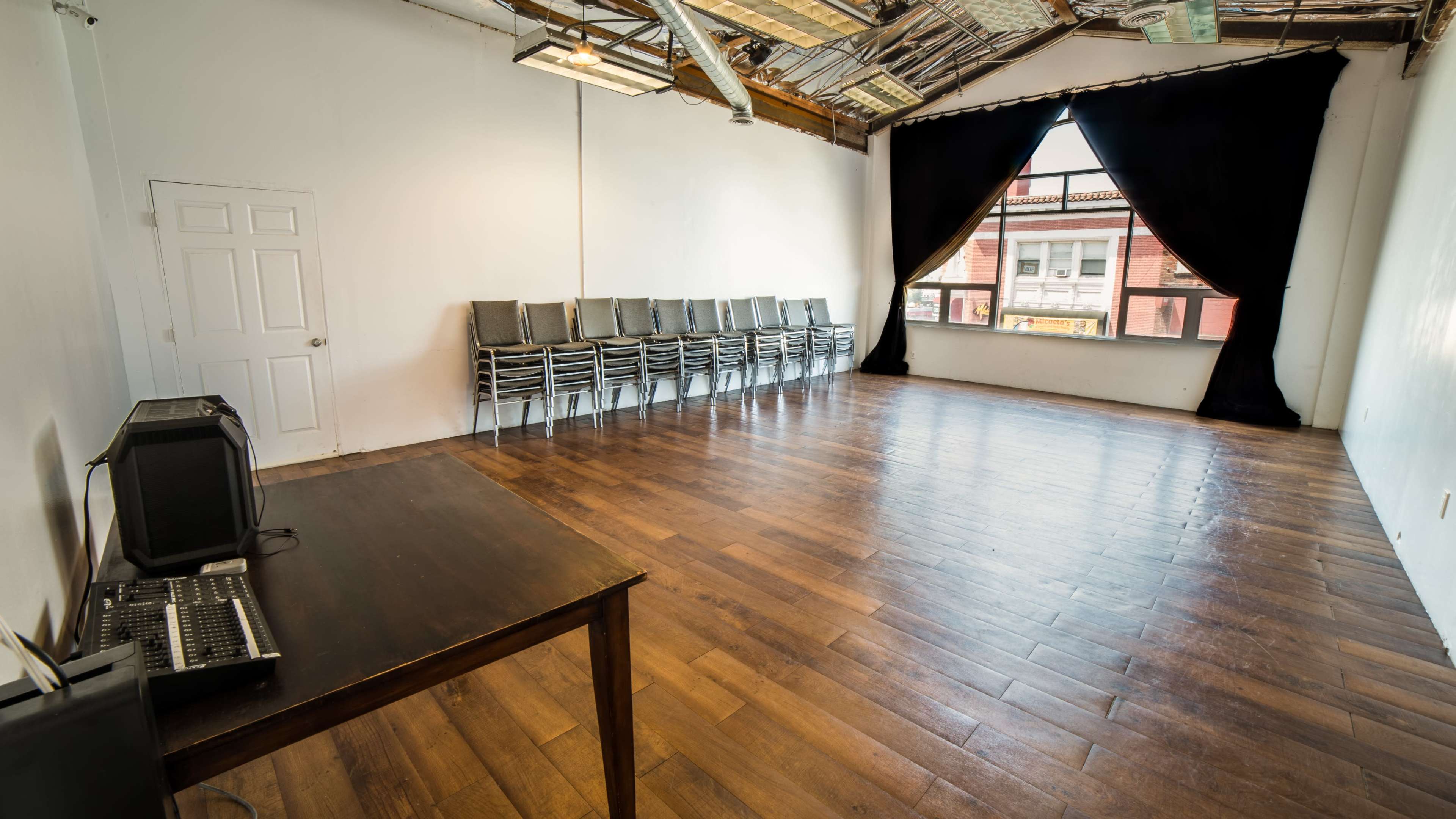 An empty room with wooden flooring, a row of stacked chairs against one wall, and a large window covered by a black curtain.