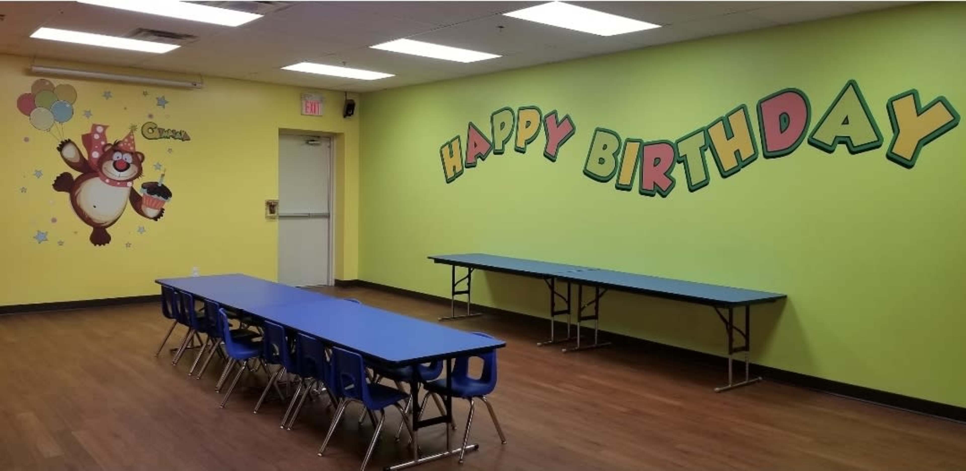 The image depicts a brightly colored birthday party room featuring yellow walls with "HAPPY BIRTHDAY" written in large letters and two blue tables arranged for guests.