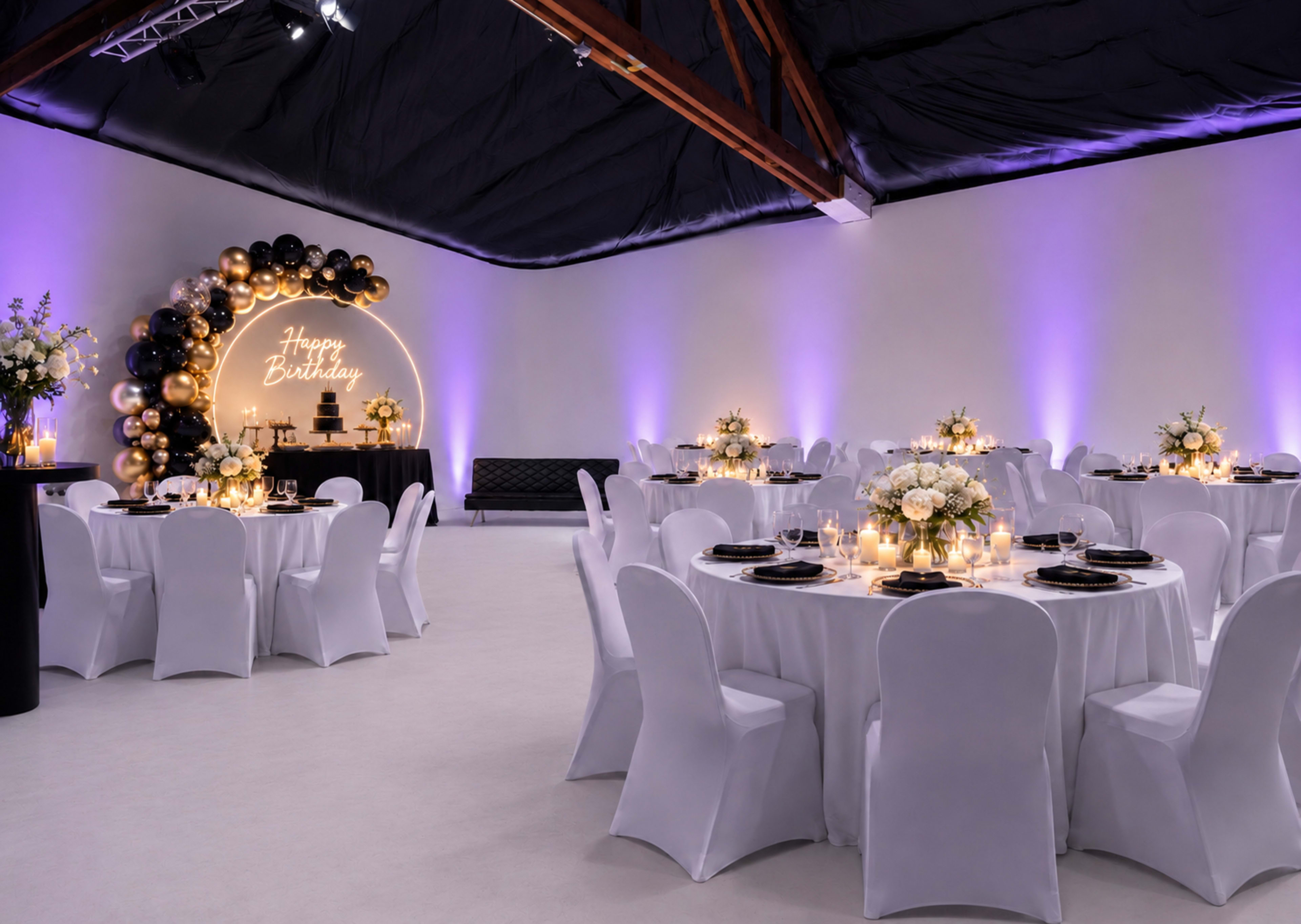 The image shows a decorated event space with round tables covered in white tablecloths, illuminated by soft lighting and featuring floral centerpieces, set up for a birthday celebration.
