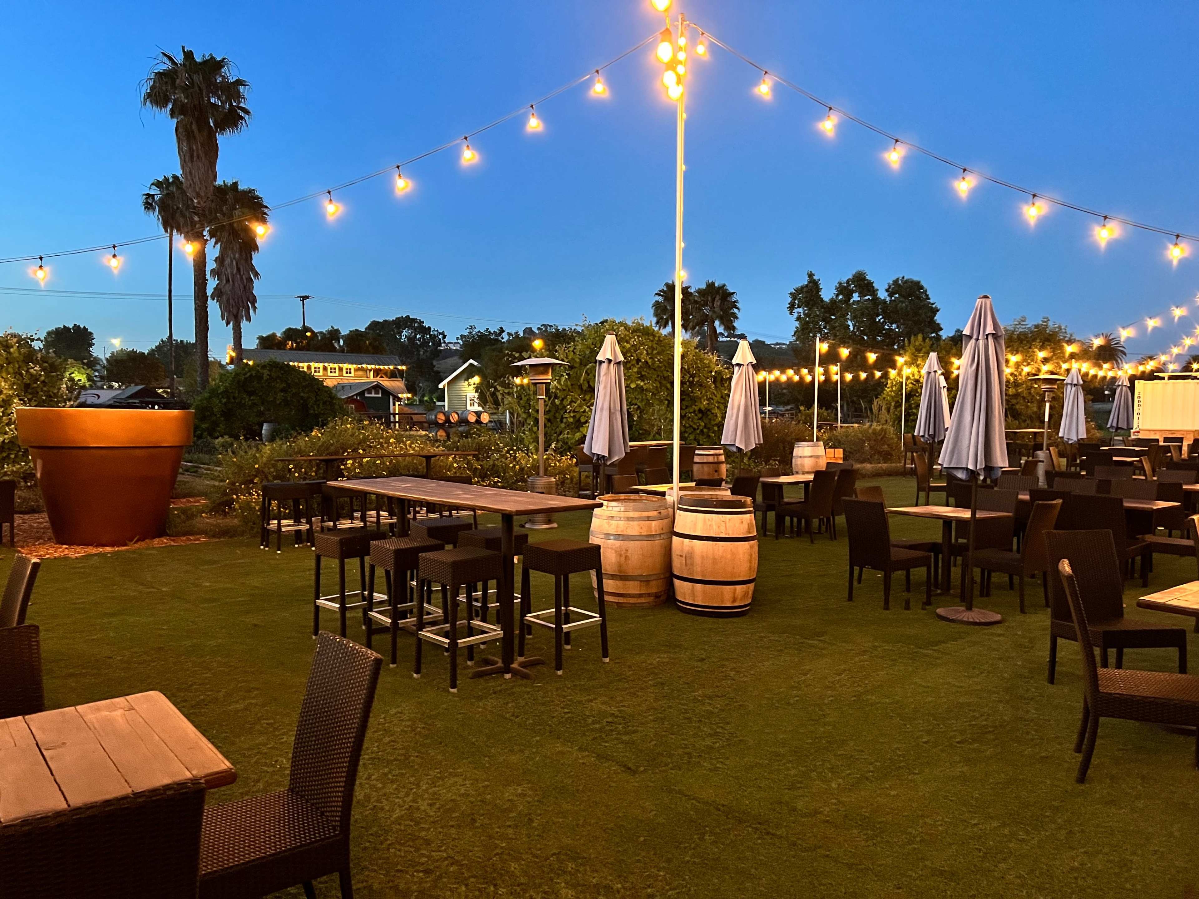 The outdoor dining area features wooden tables, high stools, and string lights, surrounded by potted palms and umbrellas at dusk.