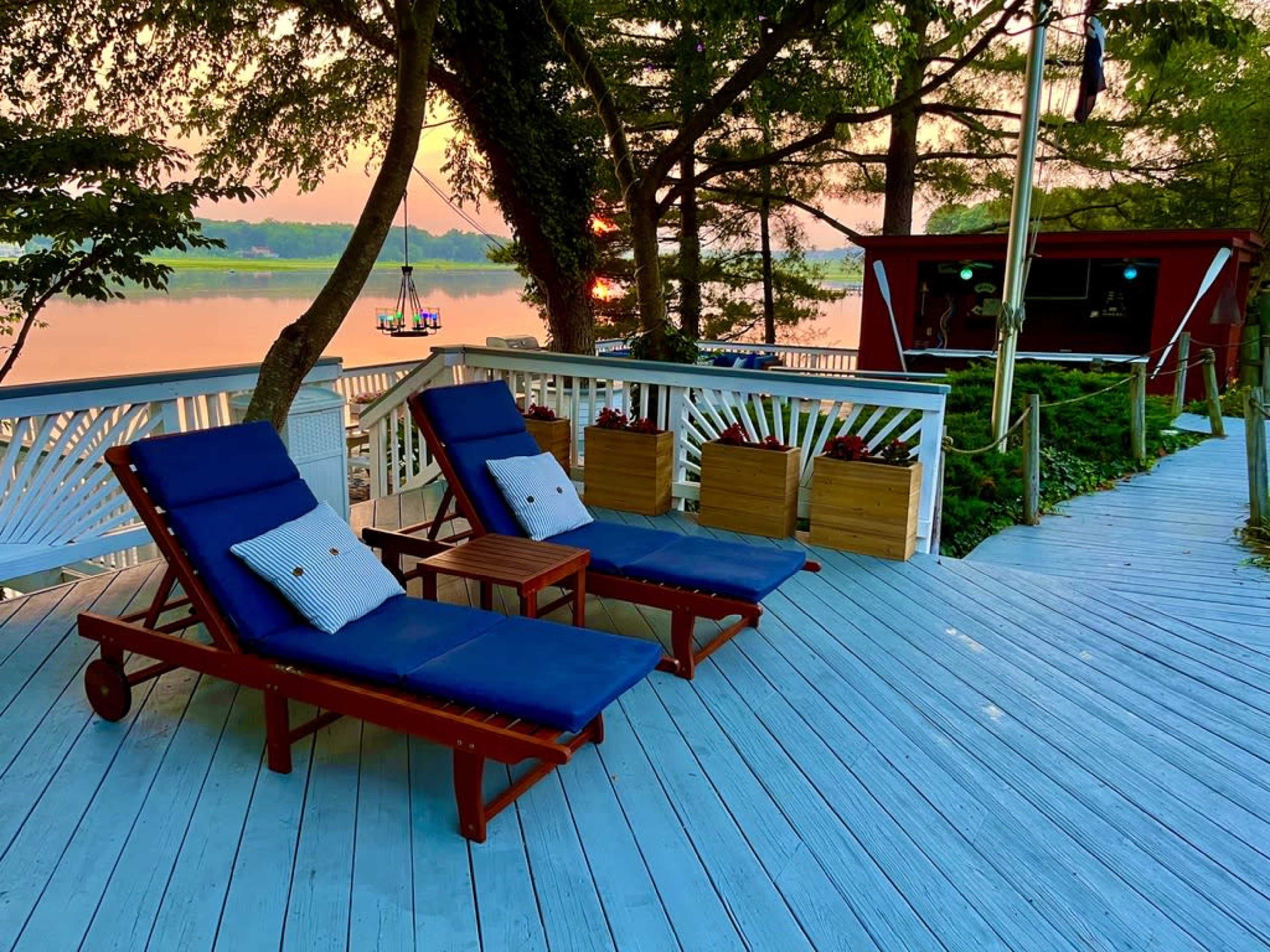 Two blue lounge chairs with cushions are positioned on a wooden deck overlooking a calm water scene at sunset.