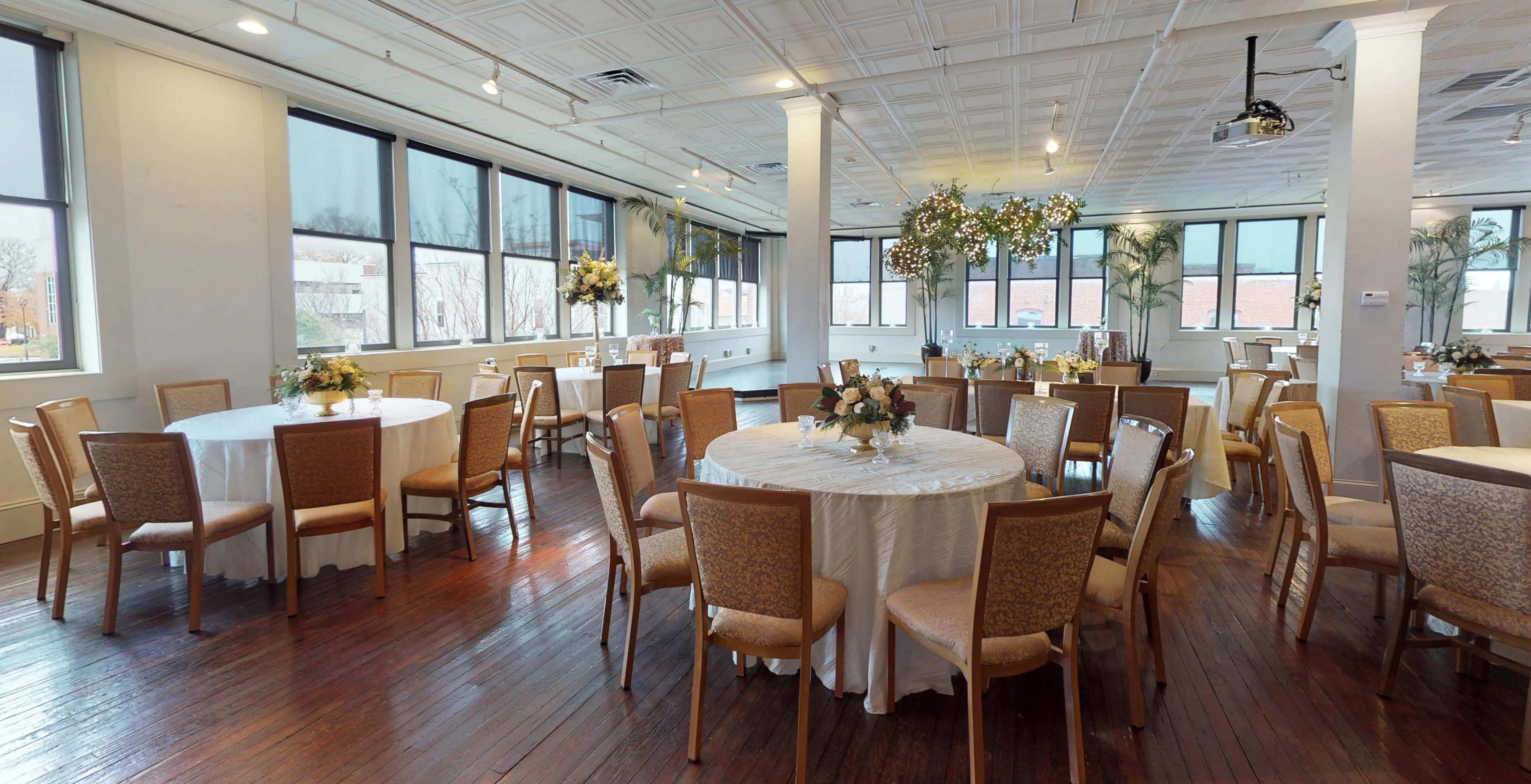 A banquet hall with several round tables, each set with floral centerpieces, arranged beneath large windows.