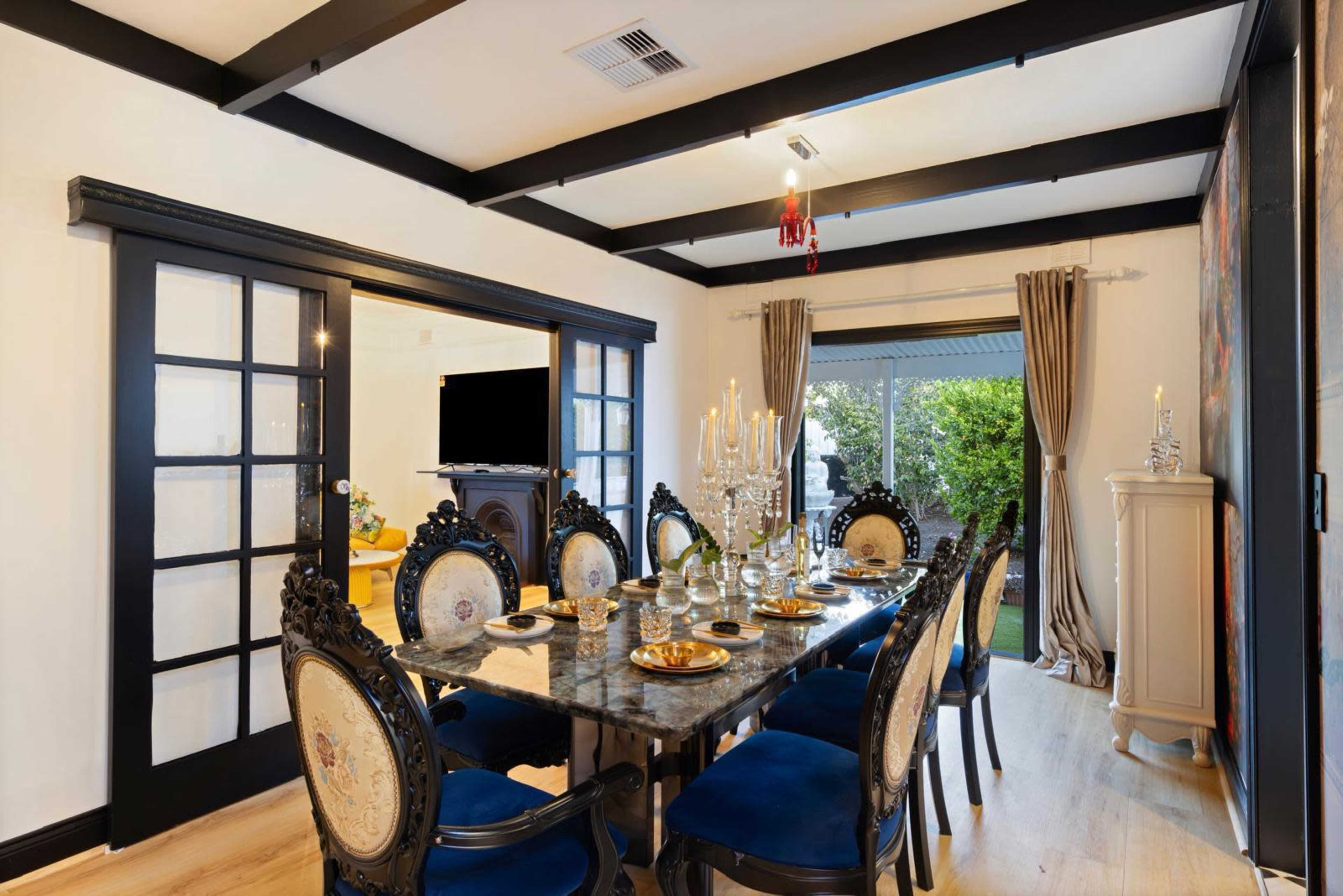 The image shows a dining room featuring a large table set for eight, surrounded by ornate chairs, with sliding glass doors that open to a view of greenery outside.
