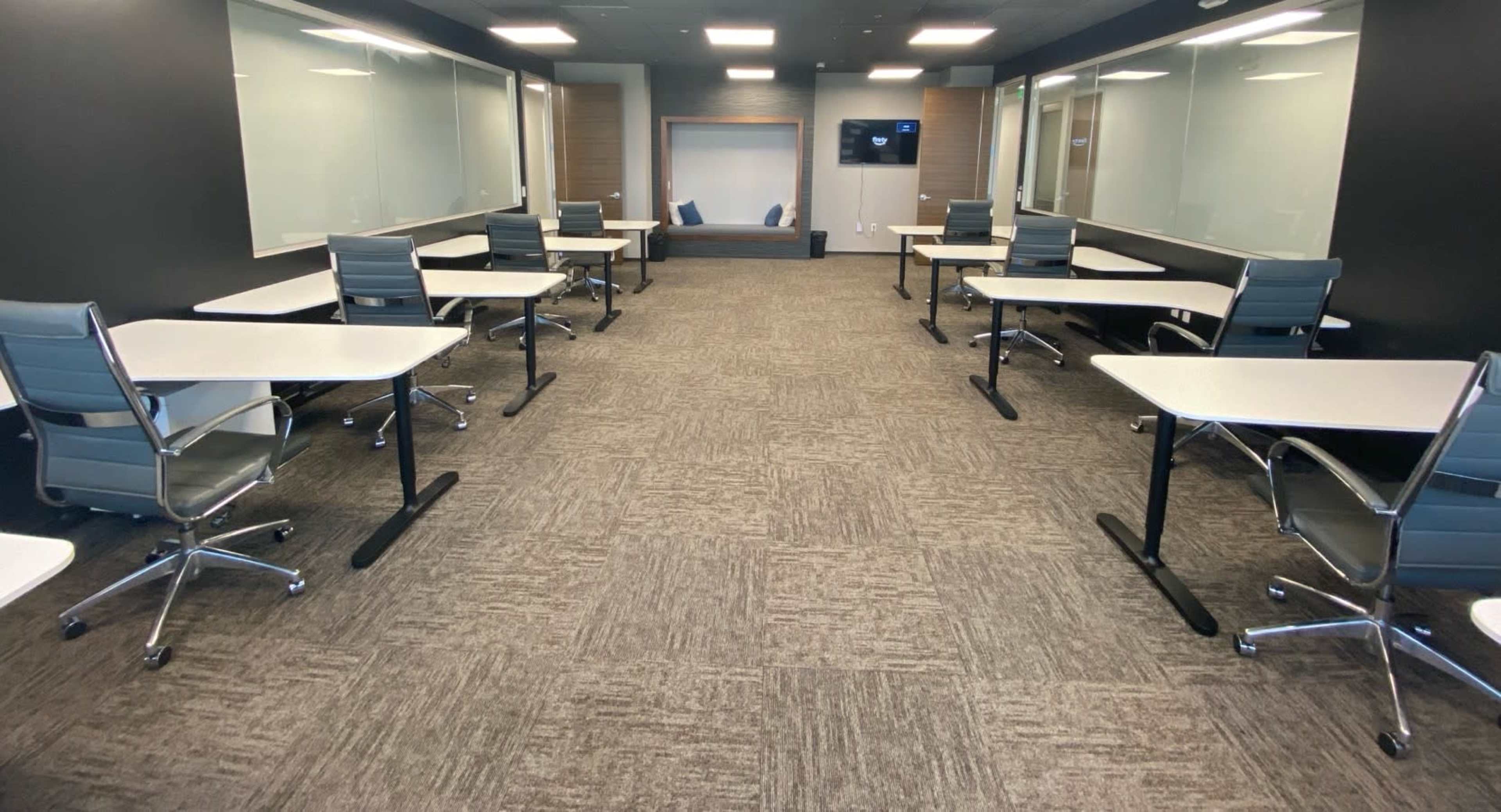 The image shows a modern conference room with several desks and chairs arranged in rows, with large windows providing natural light.