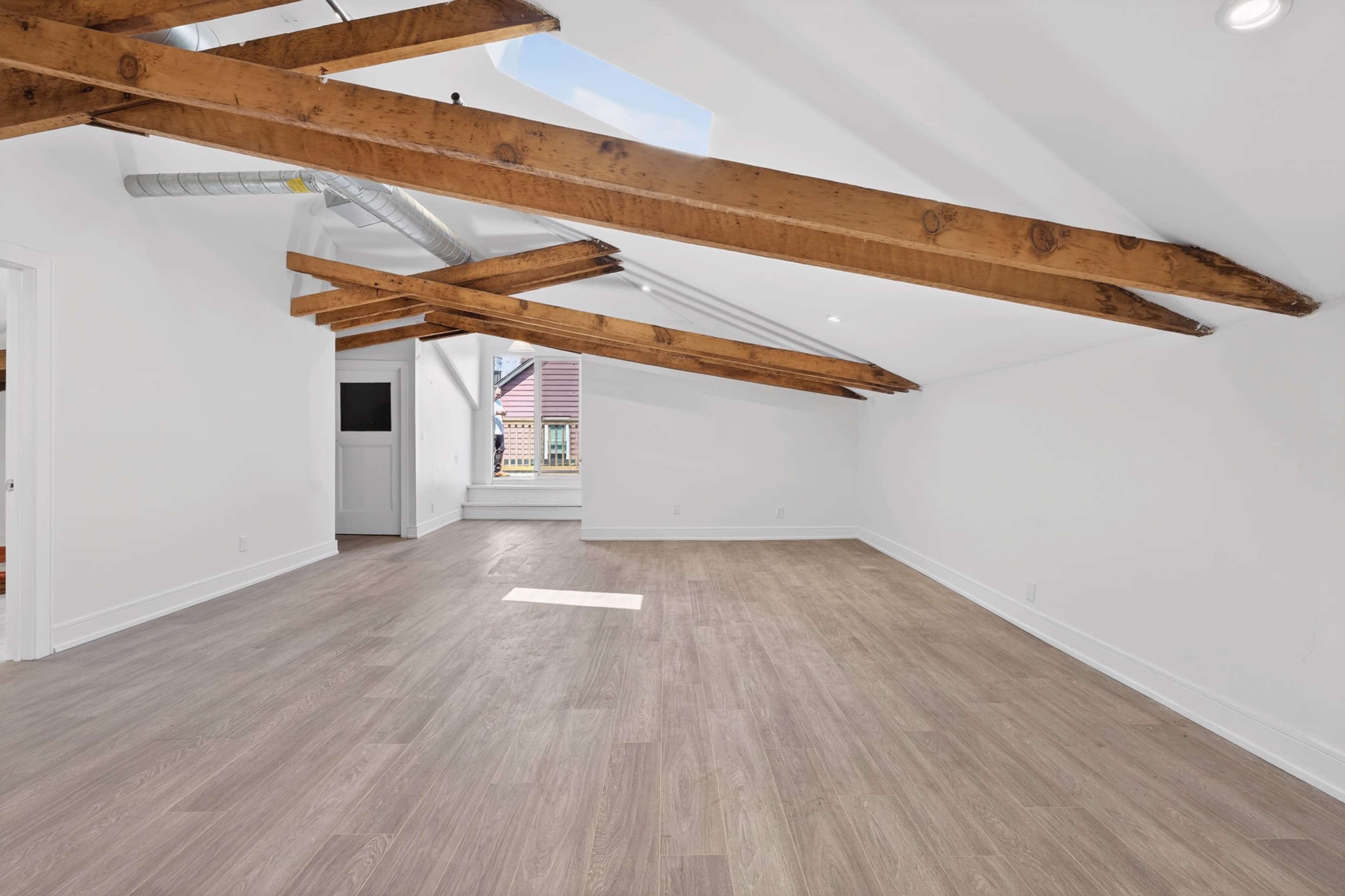 The image shows an empty, well-lit room with wooden beams on the ceiling and a skylight, featuring light-colored walls and flooring.