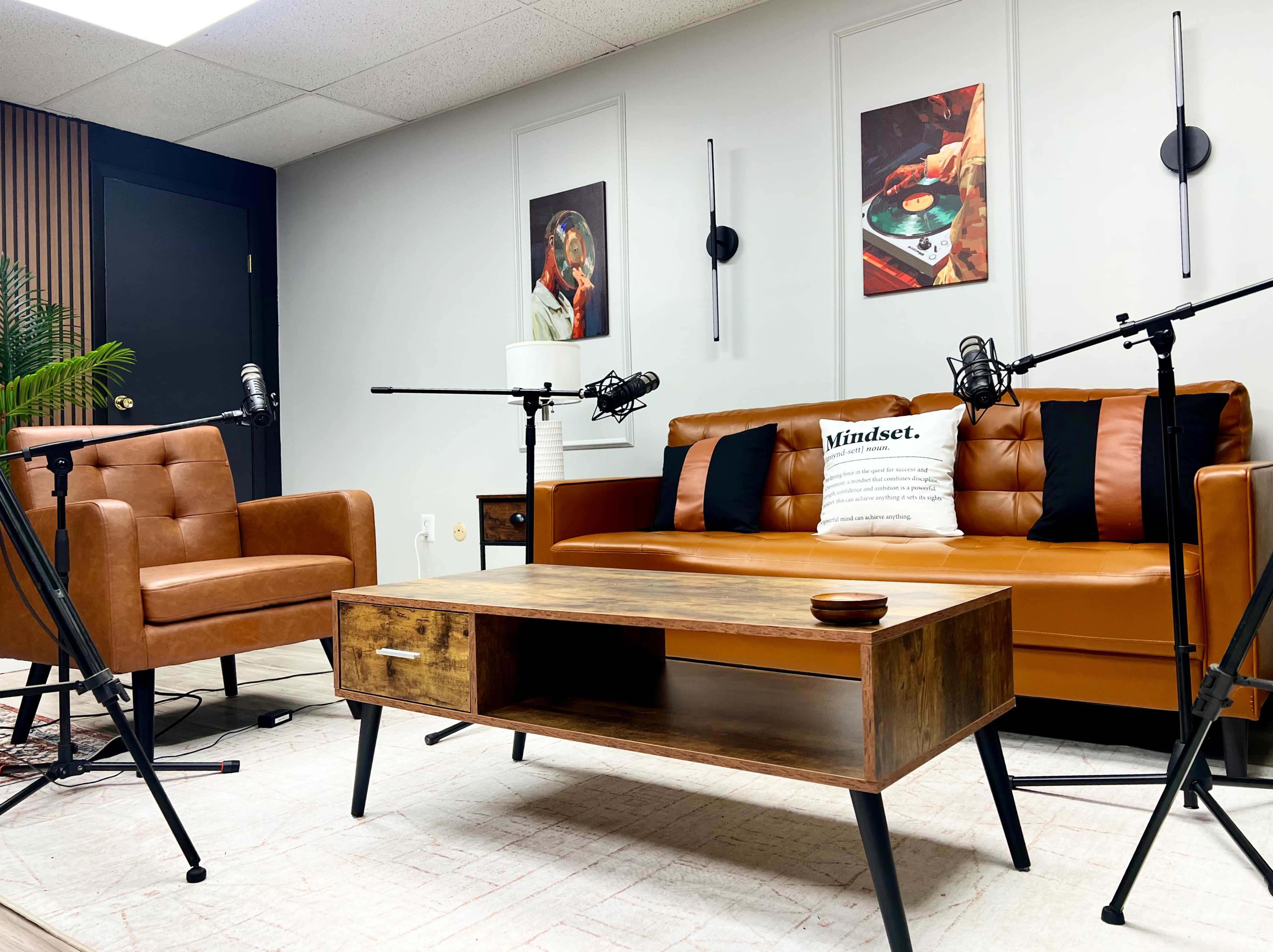 The image shows a modern podcasting studio featuring a brown leather sofa, a wooden coffee table, and two microphones positioned on stands.
