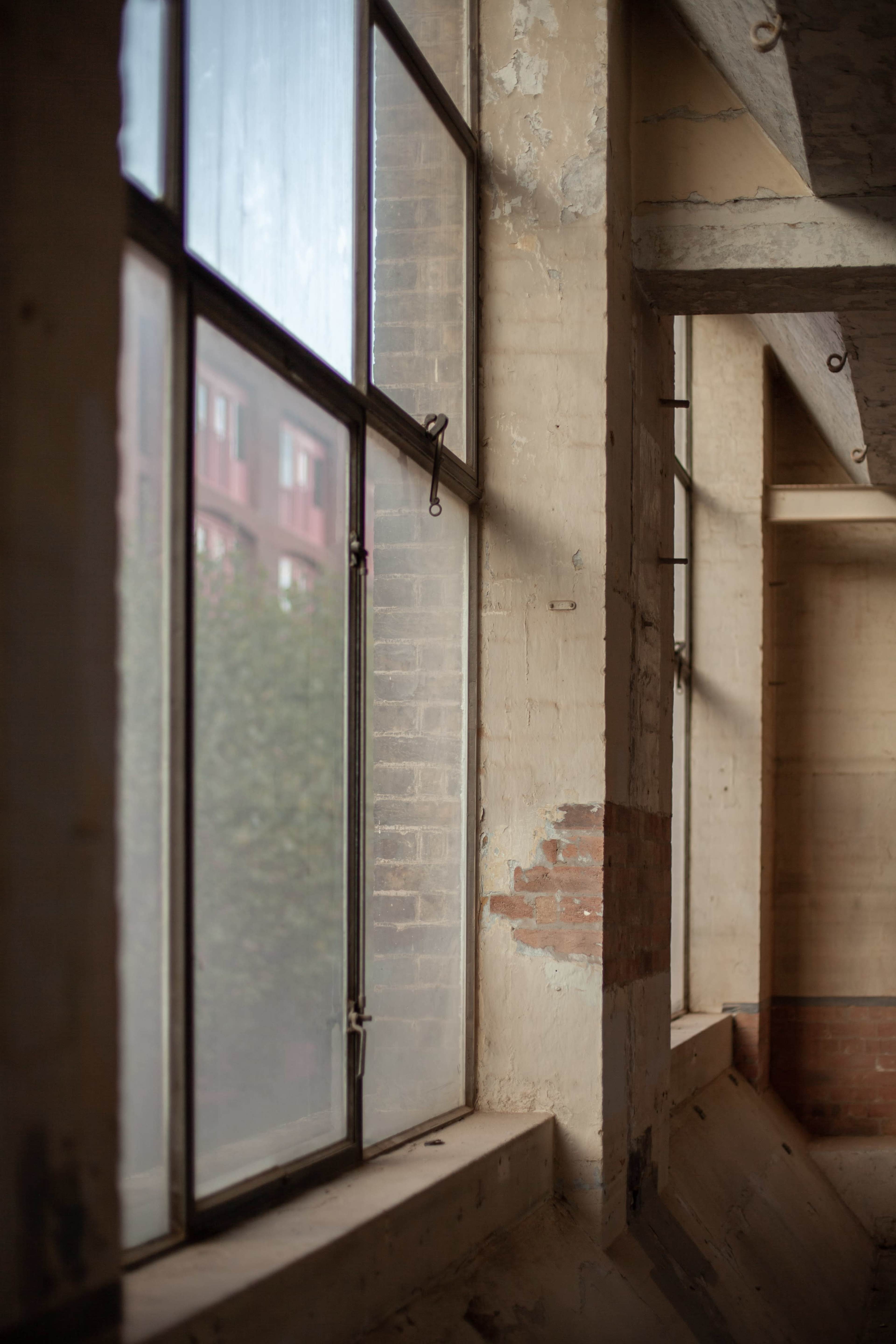 The image shows a view of large industrial windows in a partially weathered interior space with exposed brick and plaster walls.