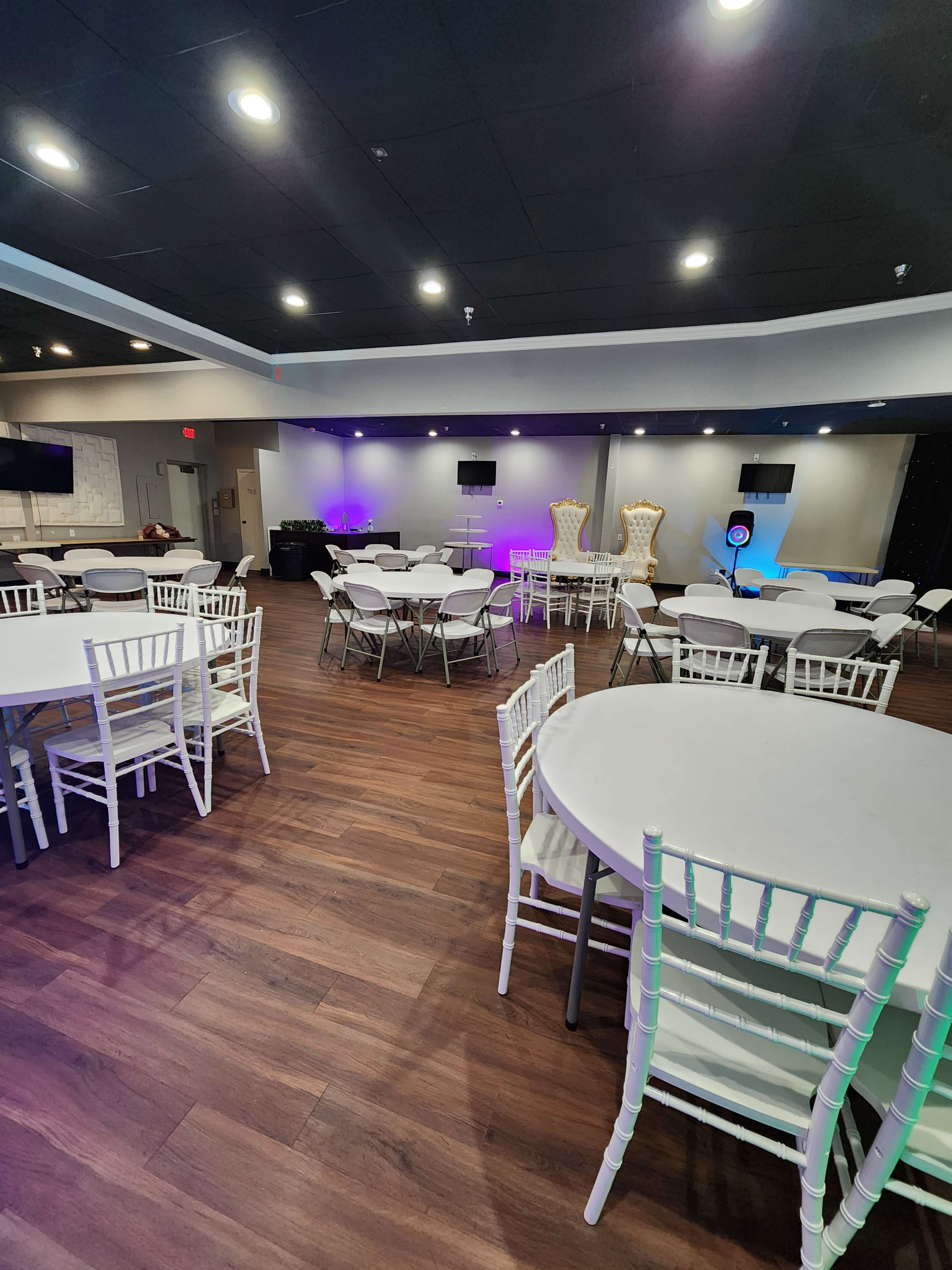 The image shows an event space with white round tables, chairs, and two large decorative chairs in the background, all under bright lighting.