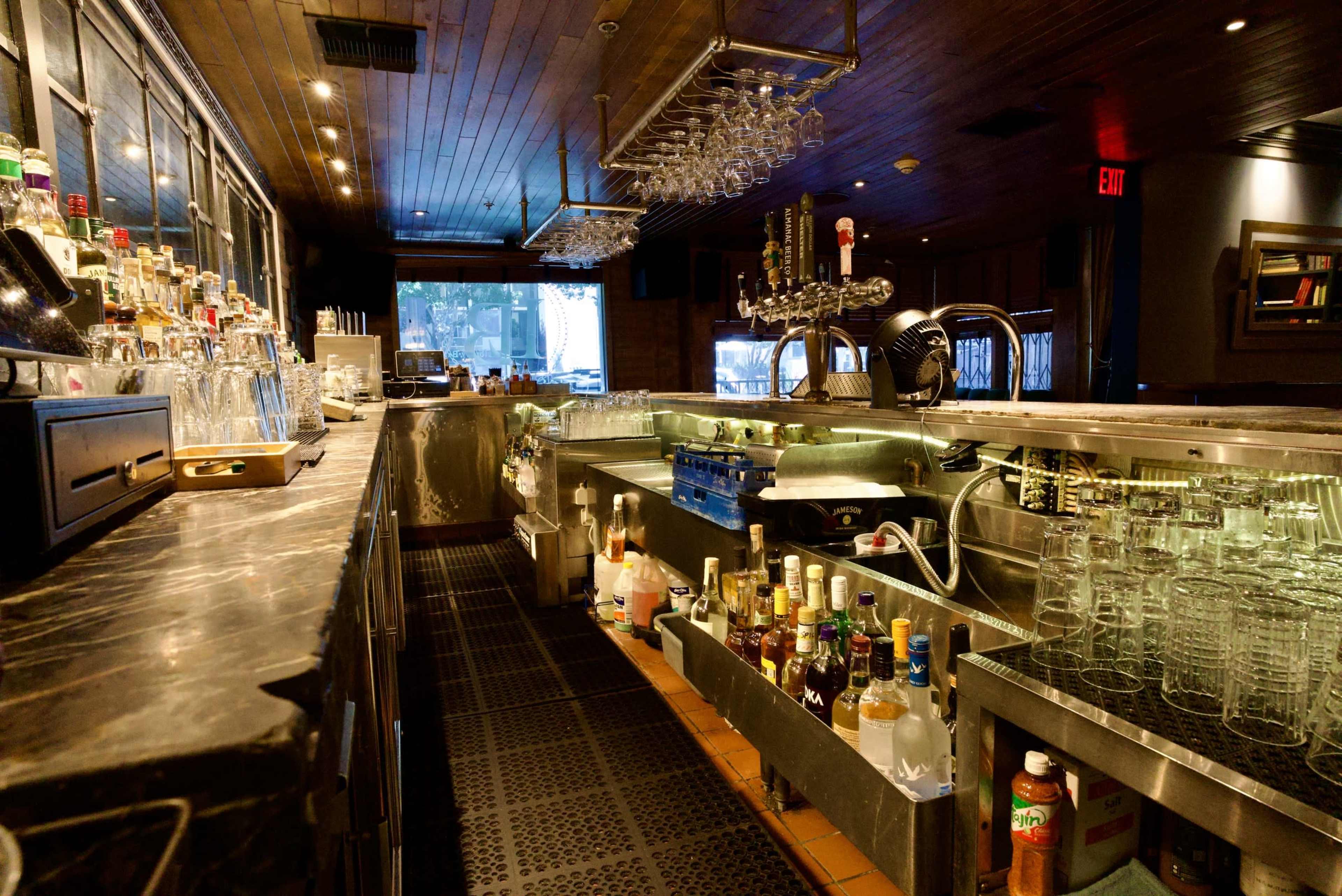 The image shows a dimly lit bar with a polished counter, shelves lined with various bottles, and glassware arranged neatly.