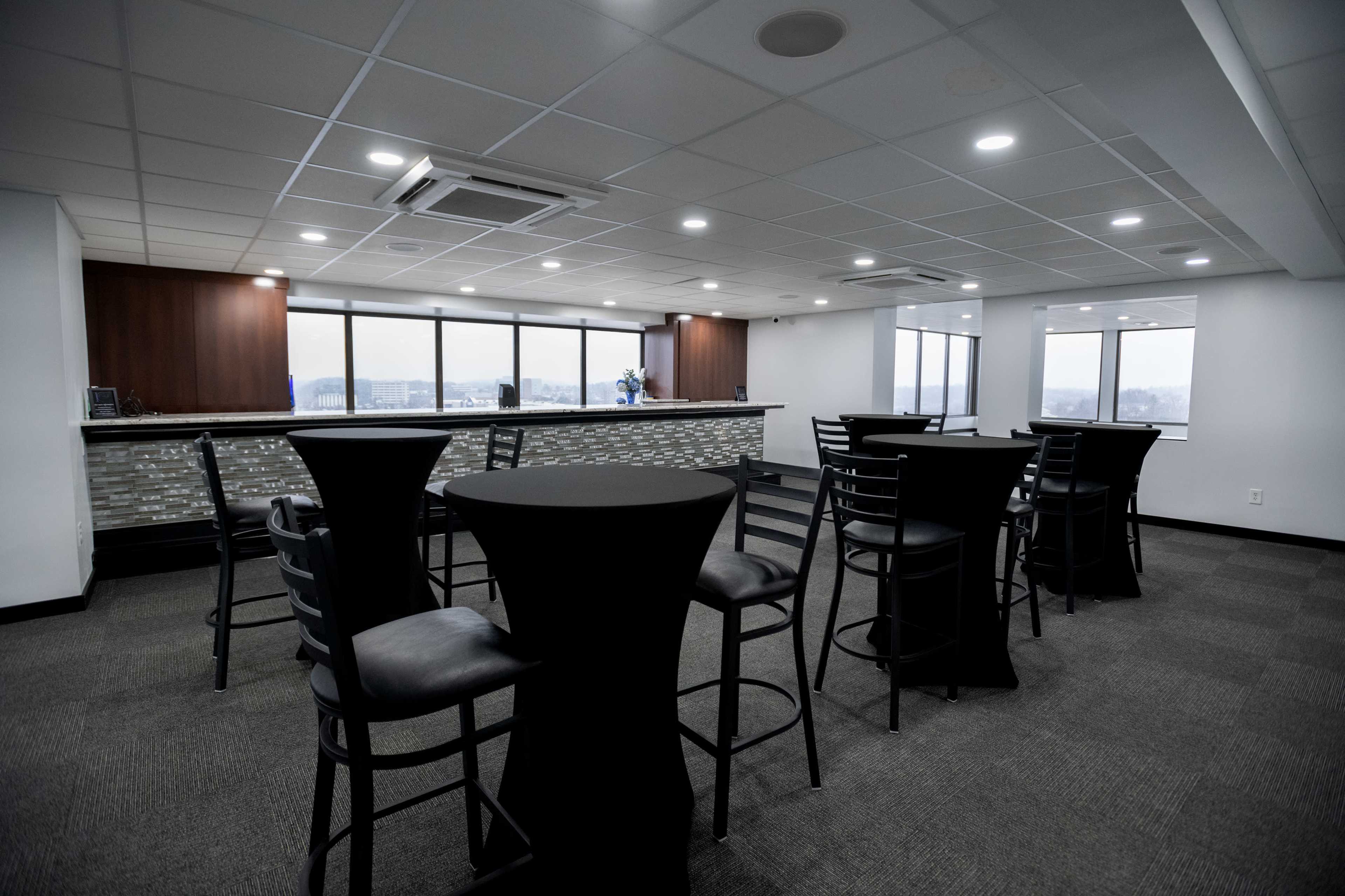 A modern interior of a bar area with several high tables and chairs, large windows, and a counter in the background.