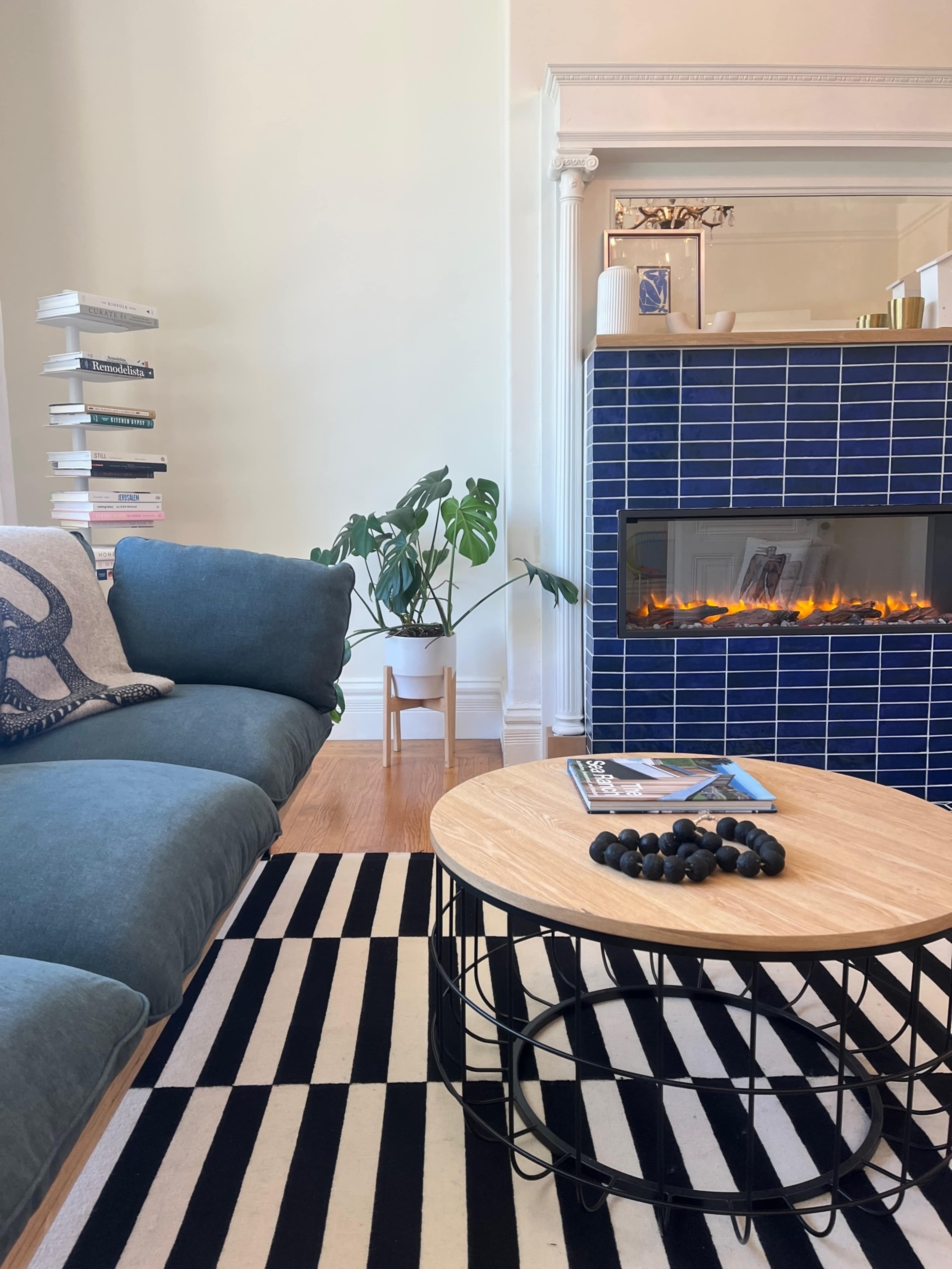 The scene features a cozy living room with a blue couch, a round wooden coffee table, a striped rug, and a blue tiled fireplace with an electric fire.