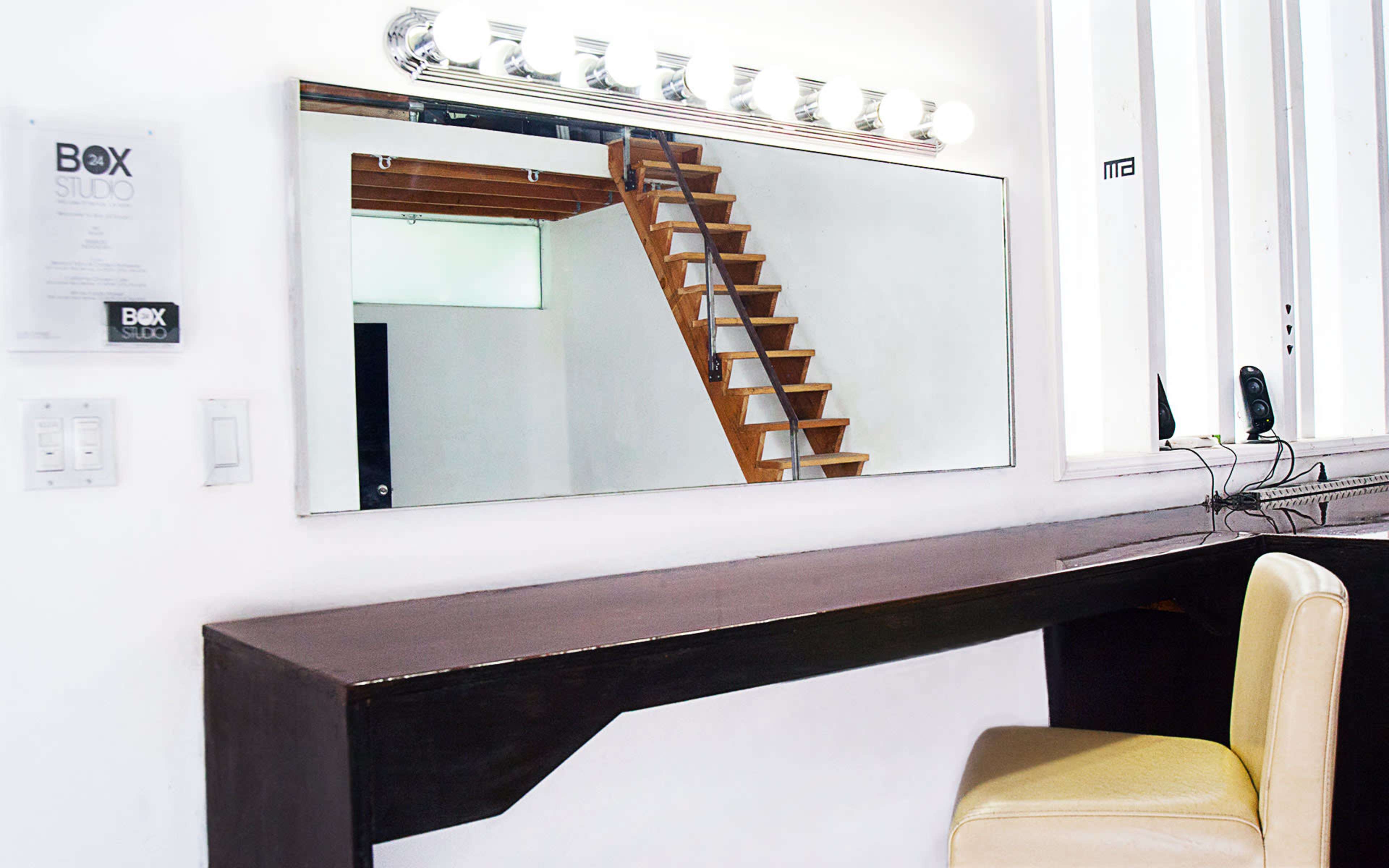 A simple vanity station with a mirror and a single beige chair is positioned against a white wall, while a wooden staircase is visible in the background.
