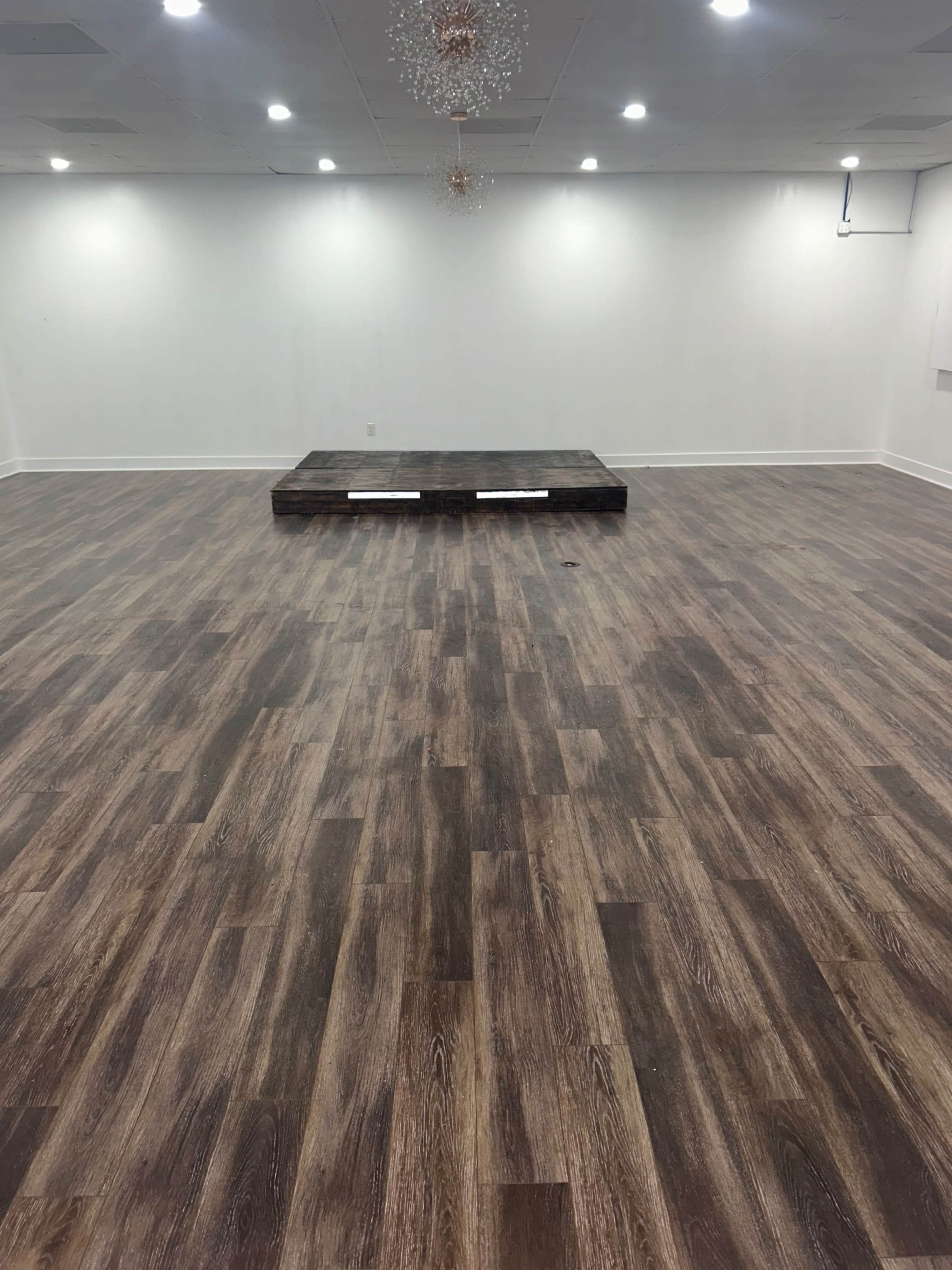 The image shows a spacious, empty room with wooden flooring and a small raised stage in the center.