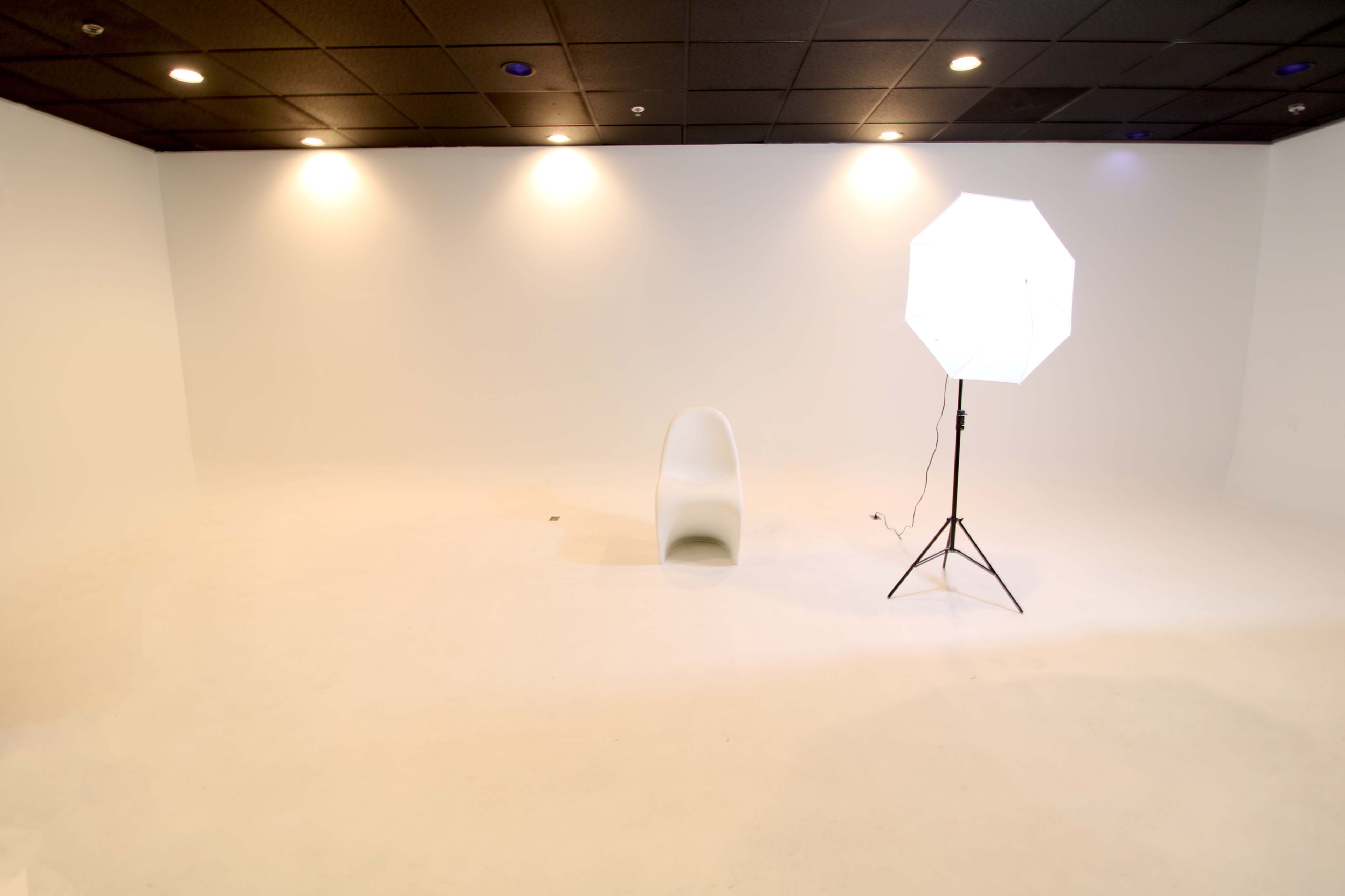 The image shows a minimalistic white studio space with a single chair and a lighting setup.