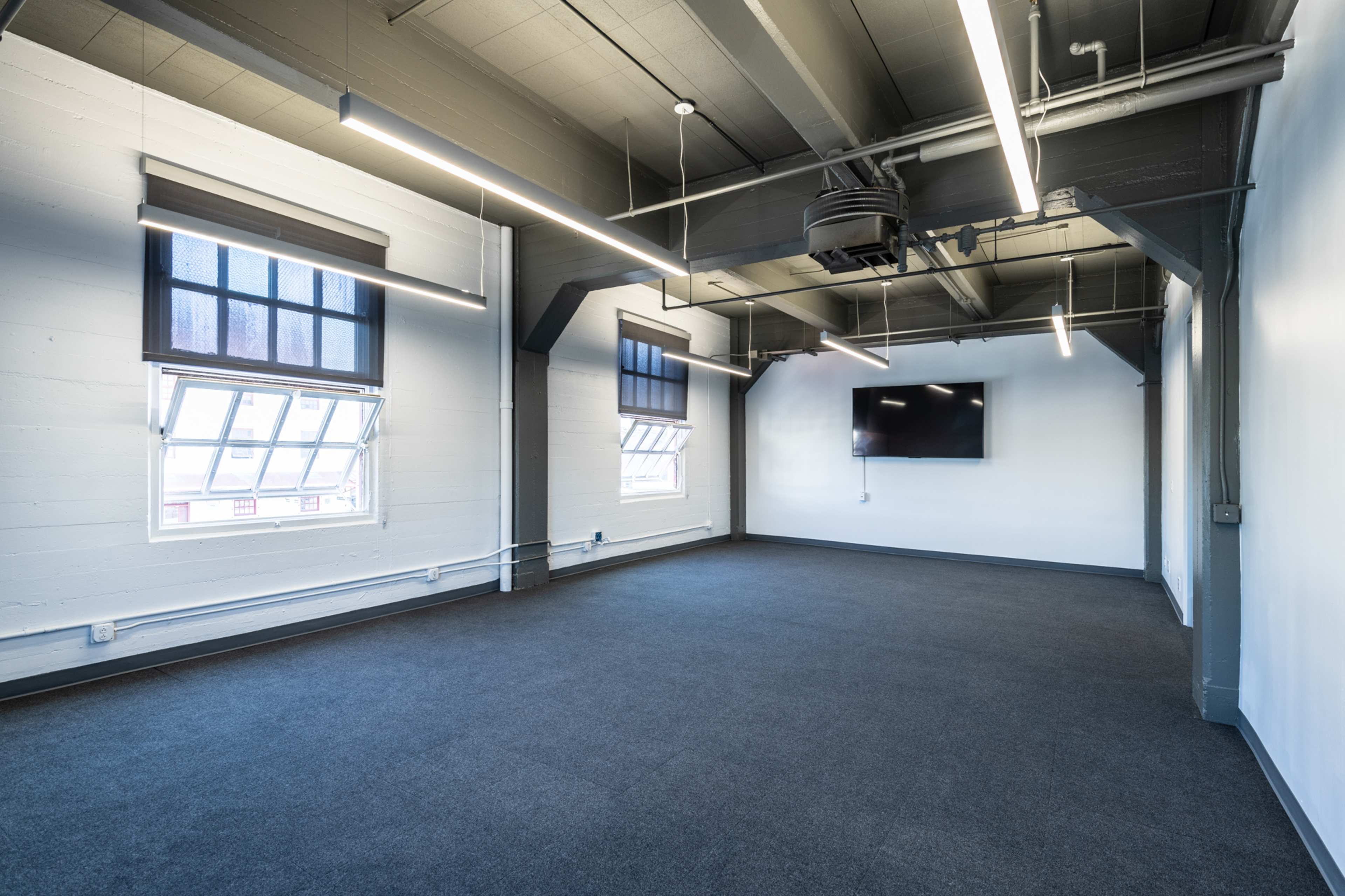 An empty room with gray carpet, large windows, and a wall-mounted television.