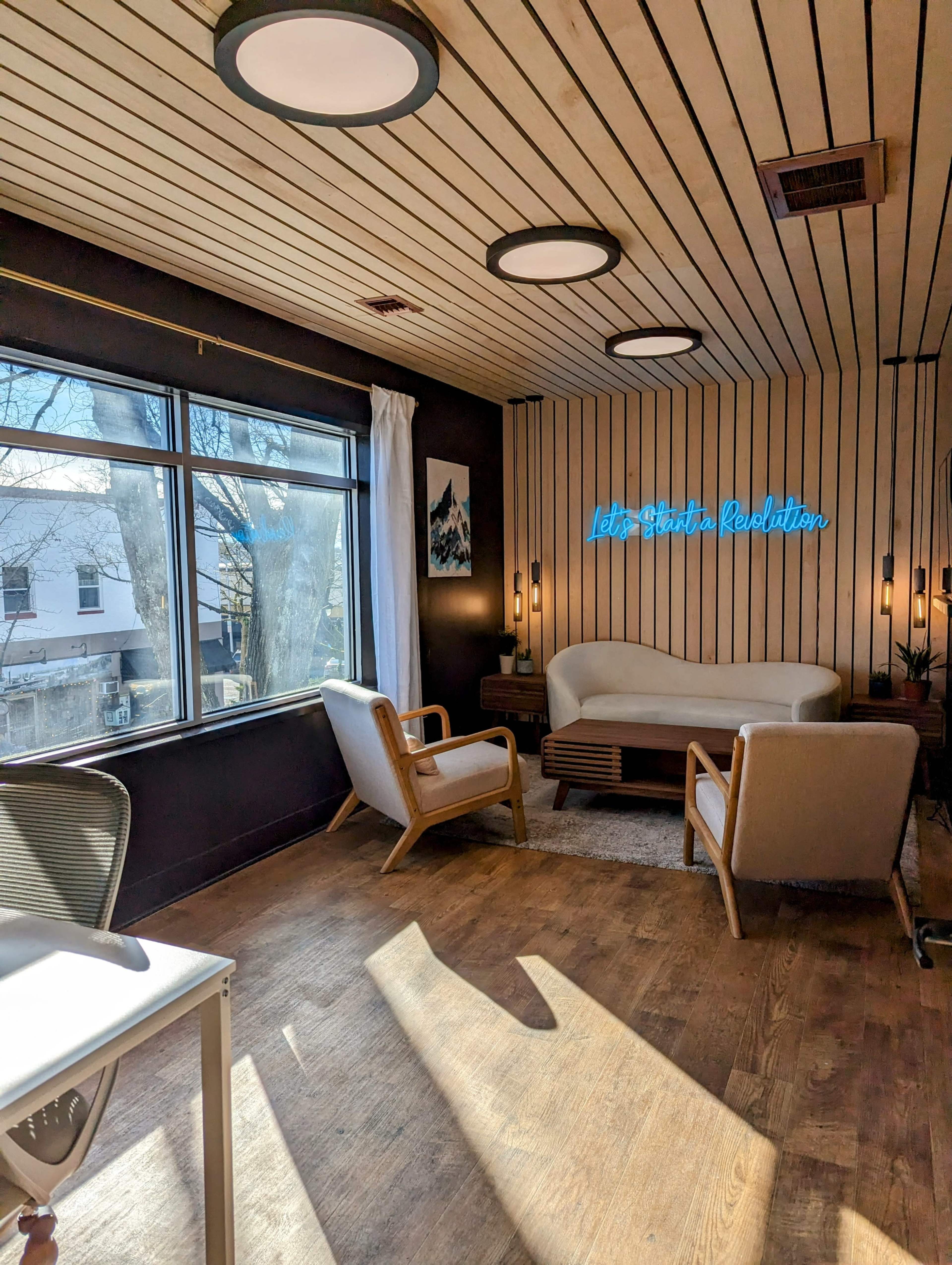 A cozy room features wooden paneling, large windows letting in natural light, and modern seating arrangements.