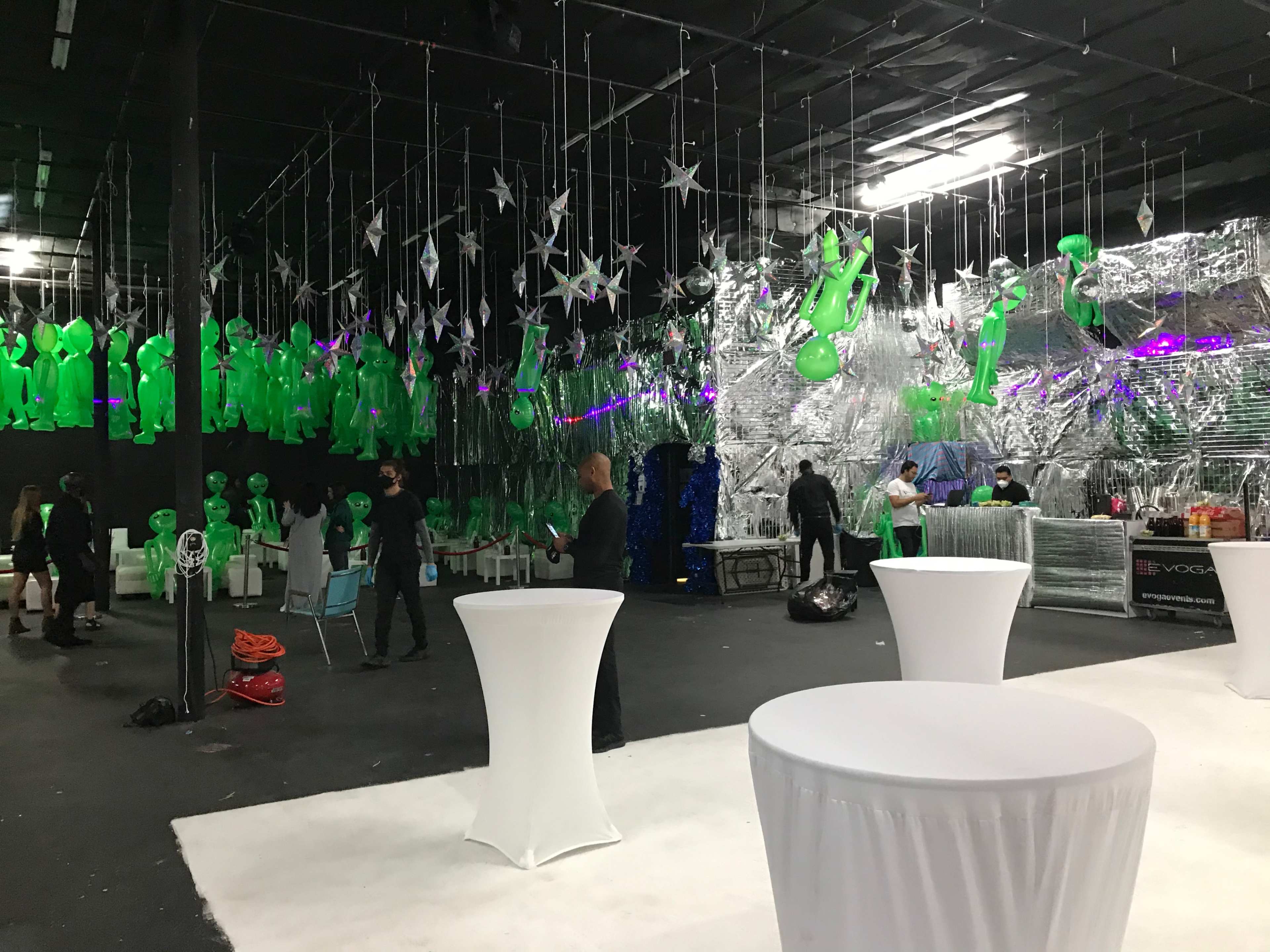 The image shows a dimly lit event space decorated with green figures and silver foil accents, featuring white cocktail tables and several people mingling.