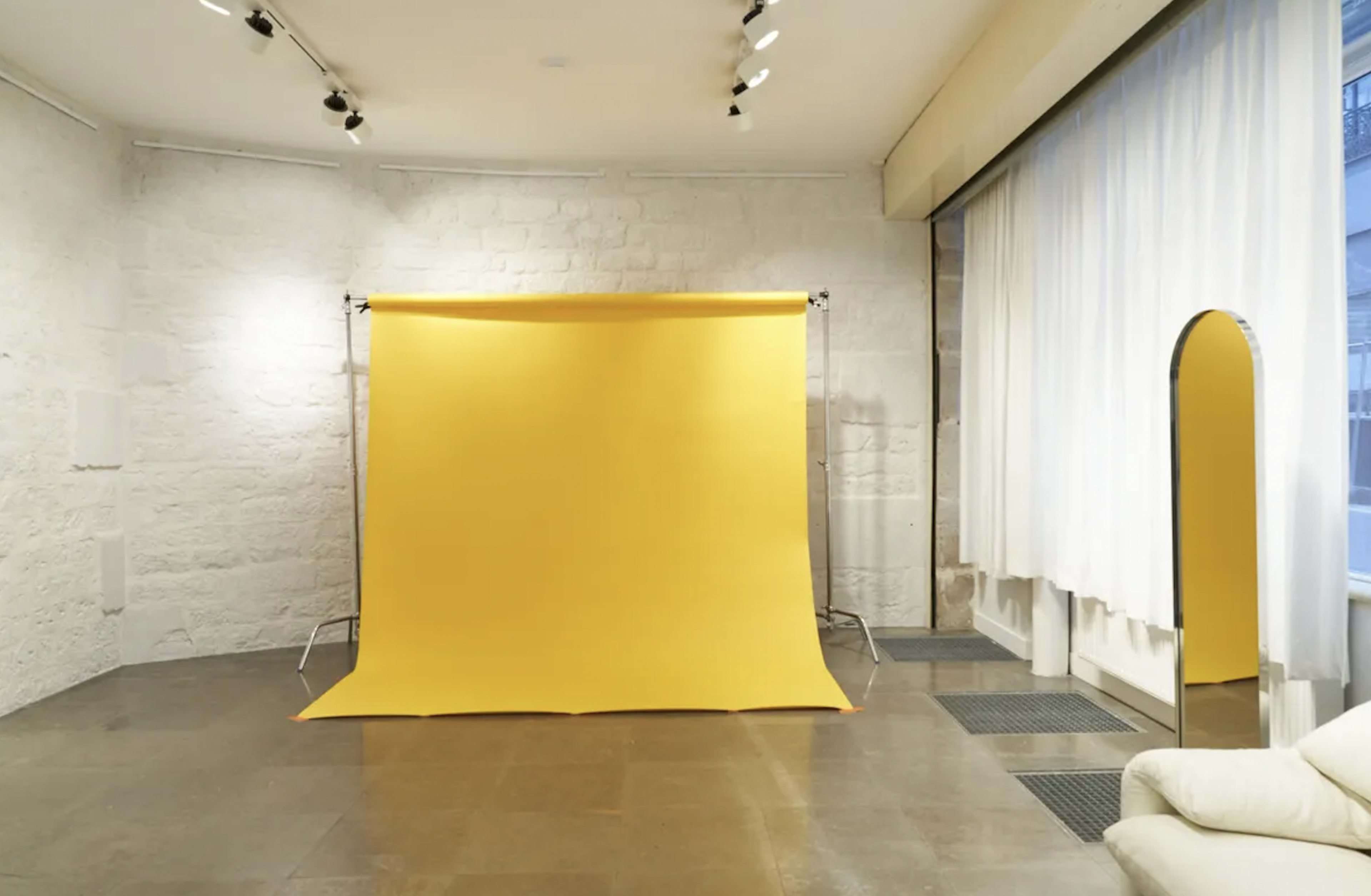 The image shows a photography studio with a yellow backdrop, a large mirror, and light-colored walls.