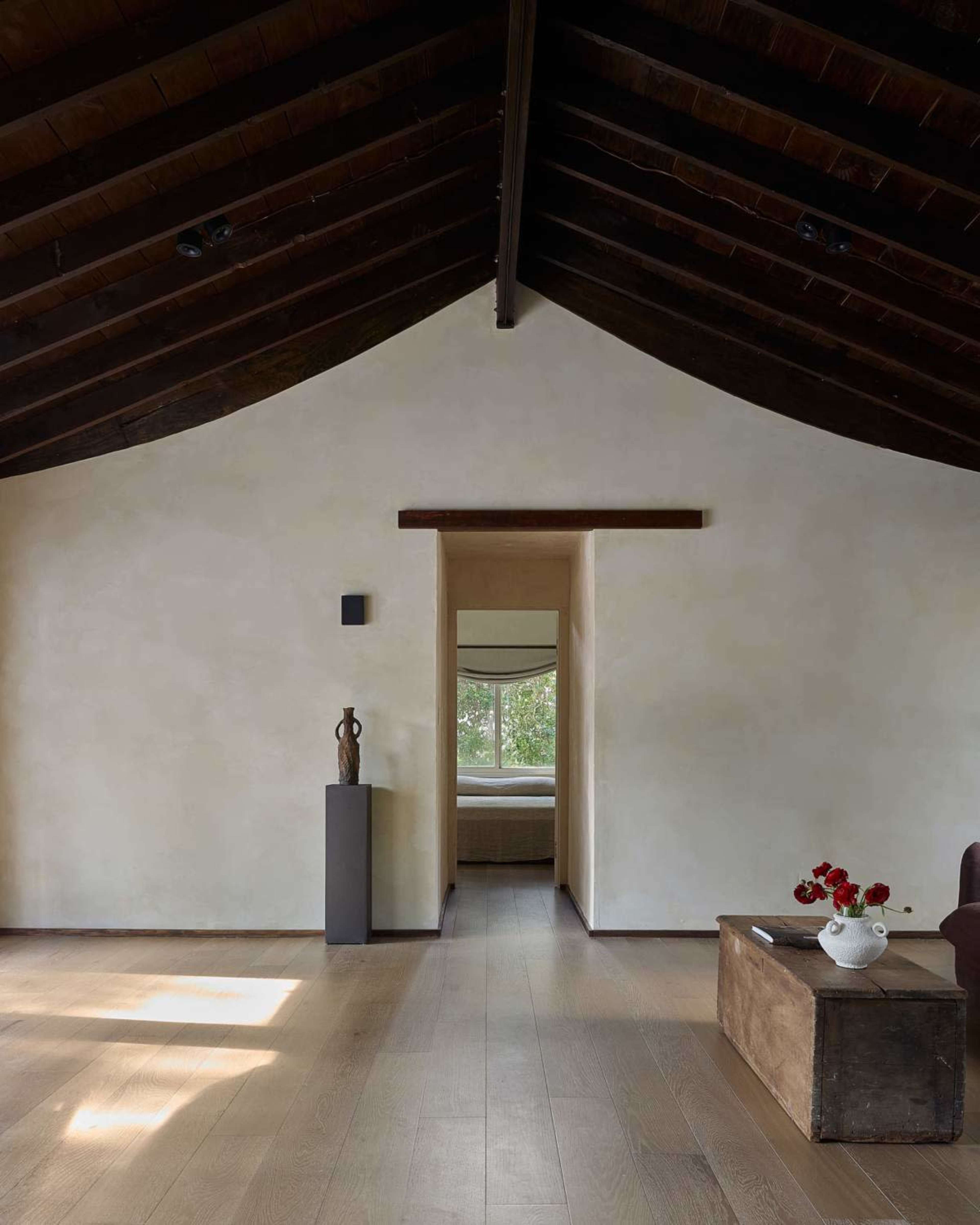 The image shows a spacious interior of a modern room with a high, sloped ceiling, light-colored walls, wooden flooring, and a view of a sunlit window framed by an architectural opening.