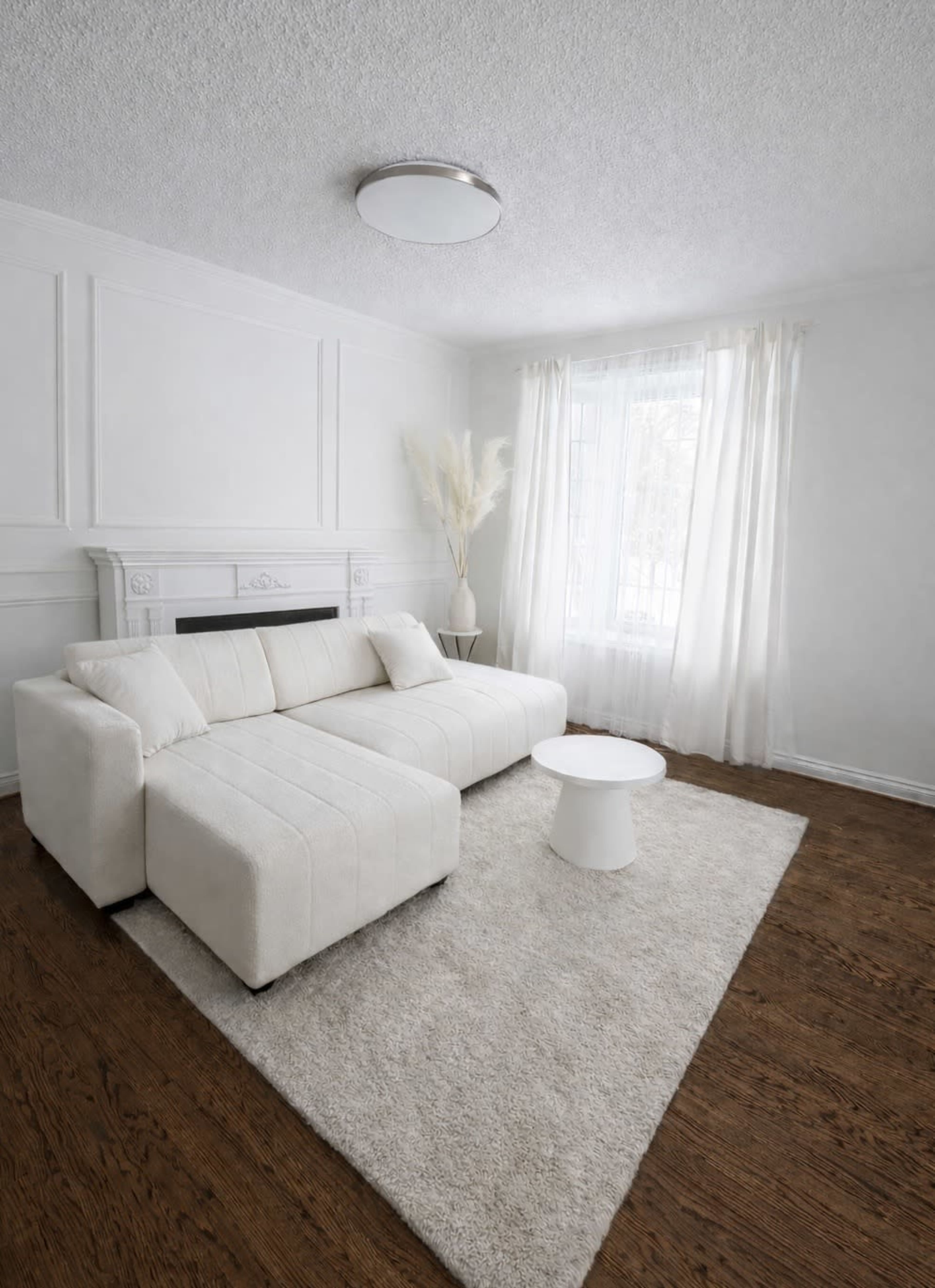 A light-filled living room features a white sectional sofa, a round coffee table, and large curtains draping a window.