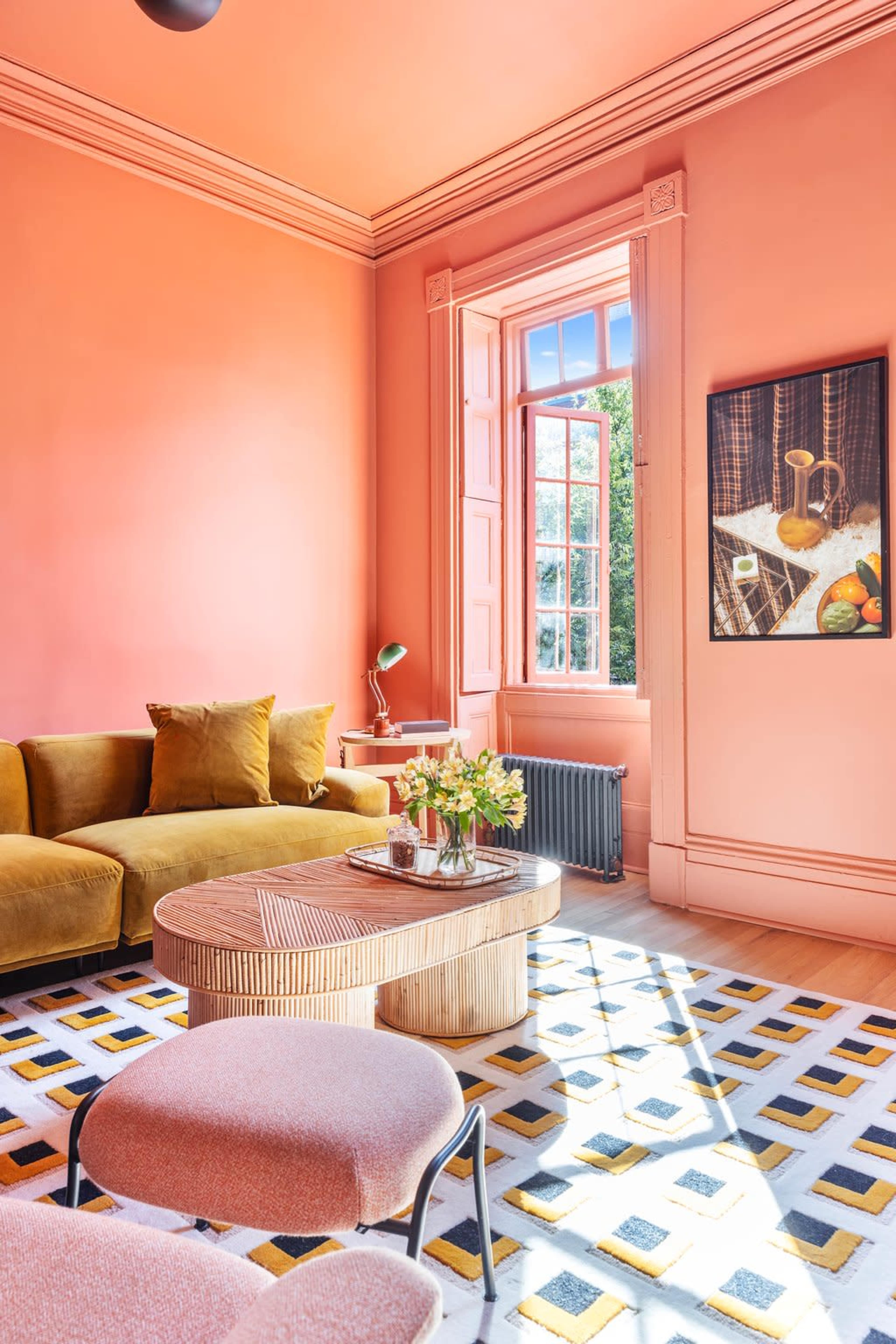 A bright living room with pink walls, a yellow sofa, a patterned rug, and a large window allowing sunlight to enter.