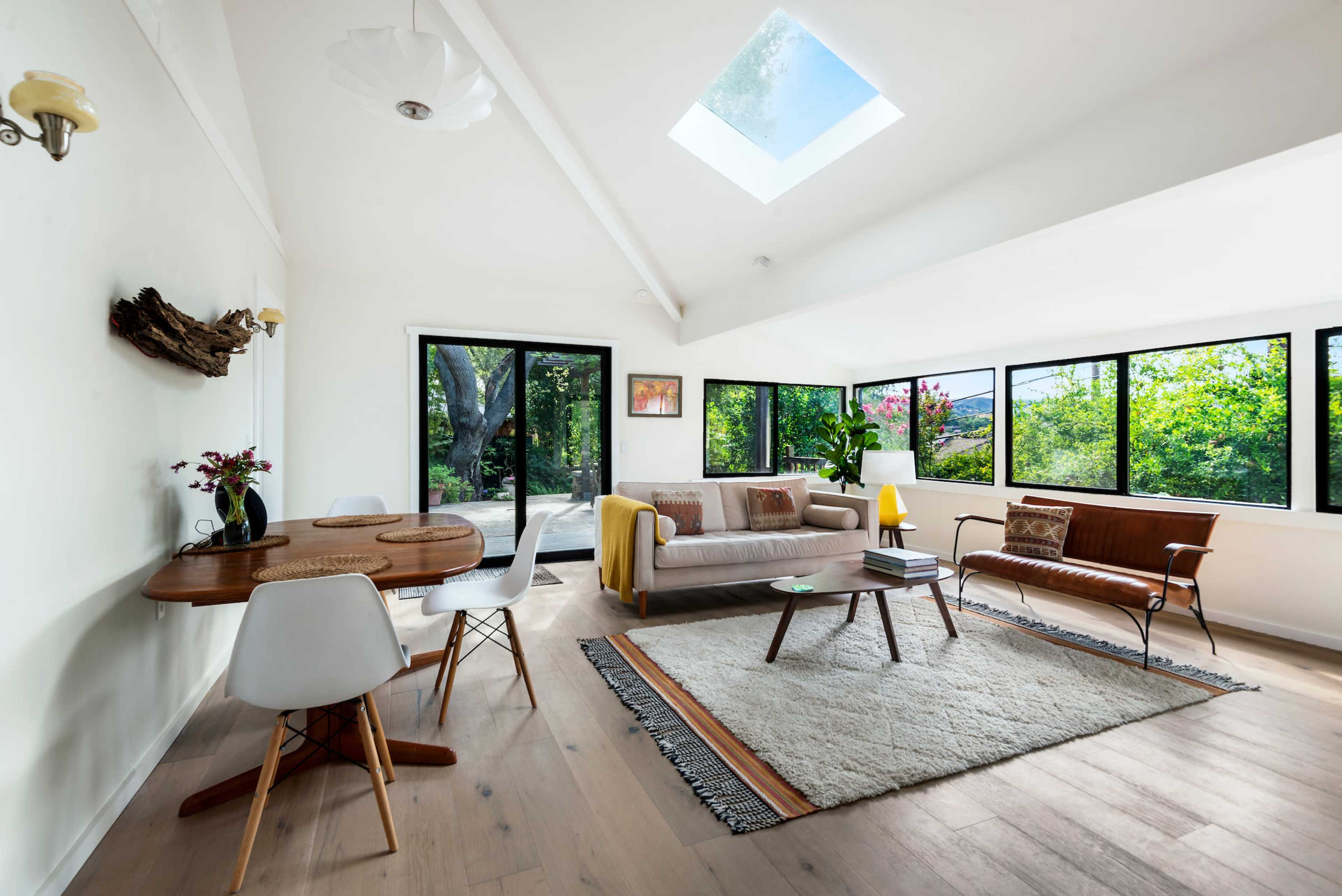 A bright living room features large windows overlooking greenery, with a sofa, dining table, and a patterned rug on a wooden floor.