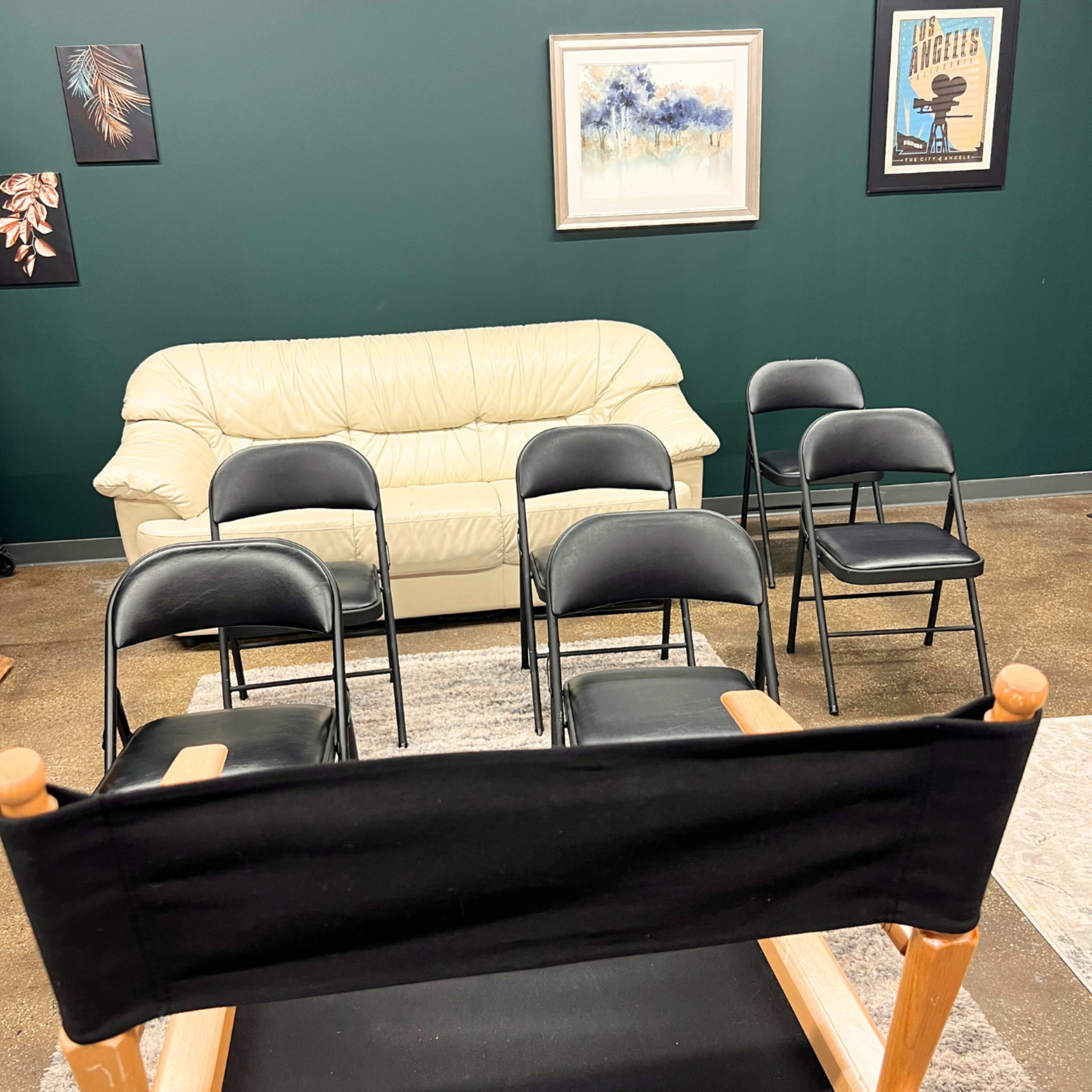 A cream-colored couch facing a row of five black folding chairs in a room with dark green walls and framed artwork.