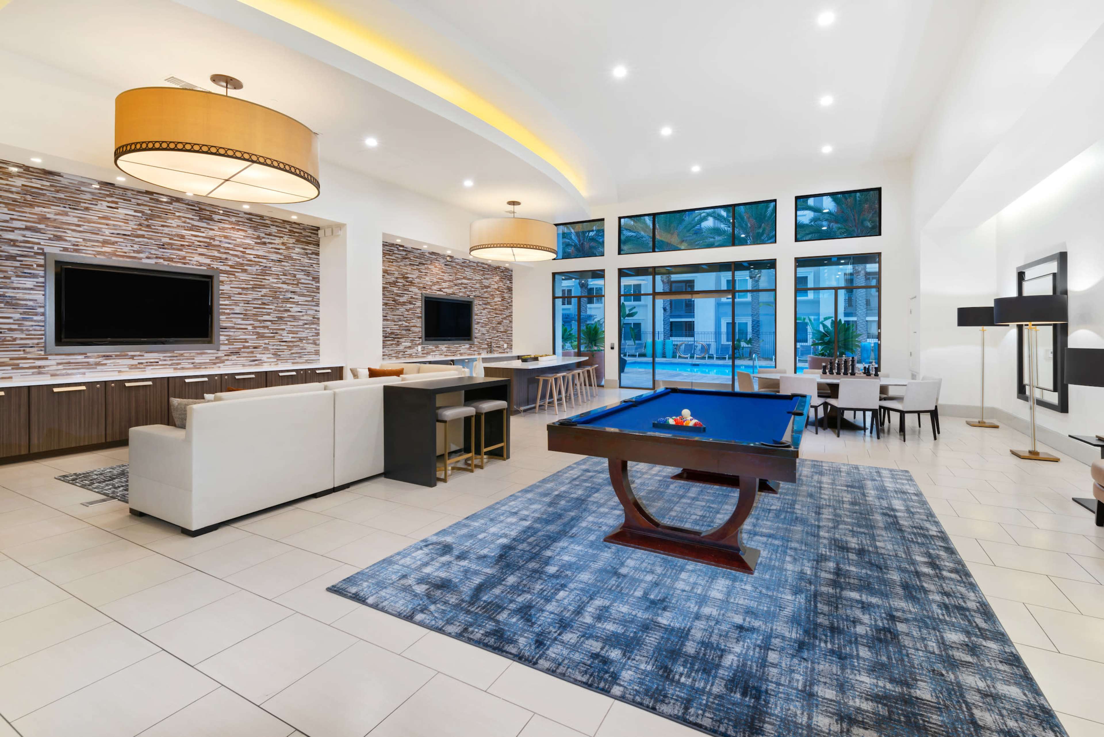 A modern interior recreational room featuring a blue pool table, seating areas, and large windows allowing natural light.