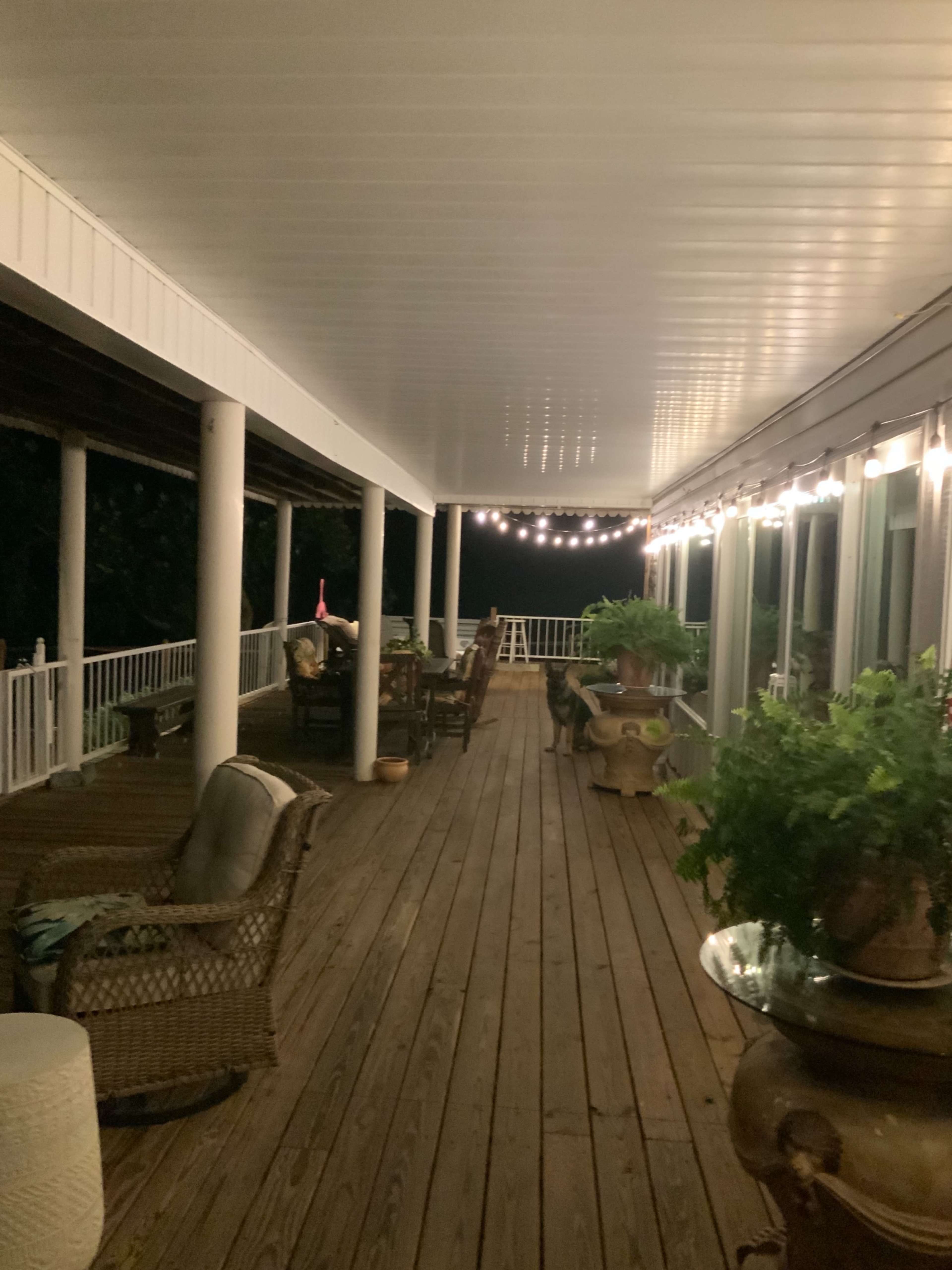 A covered wooden porch illuminated by string lights, with seating areas and potted plants along the railing.