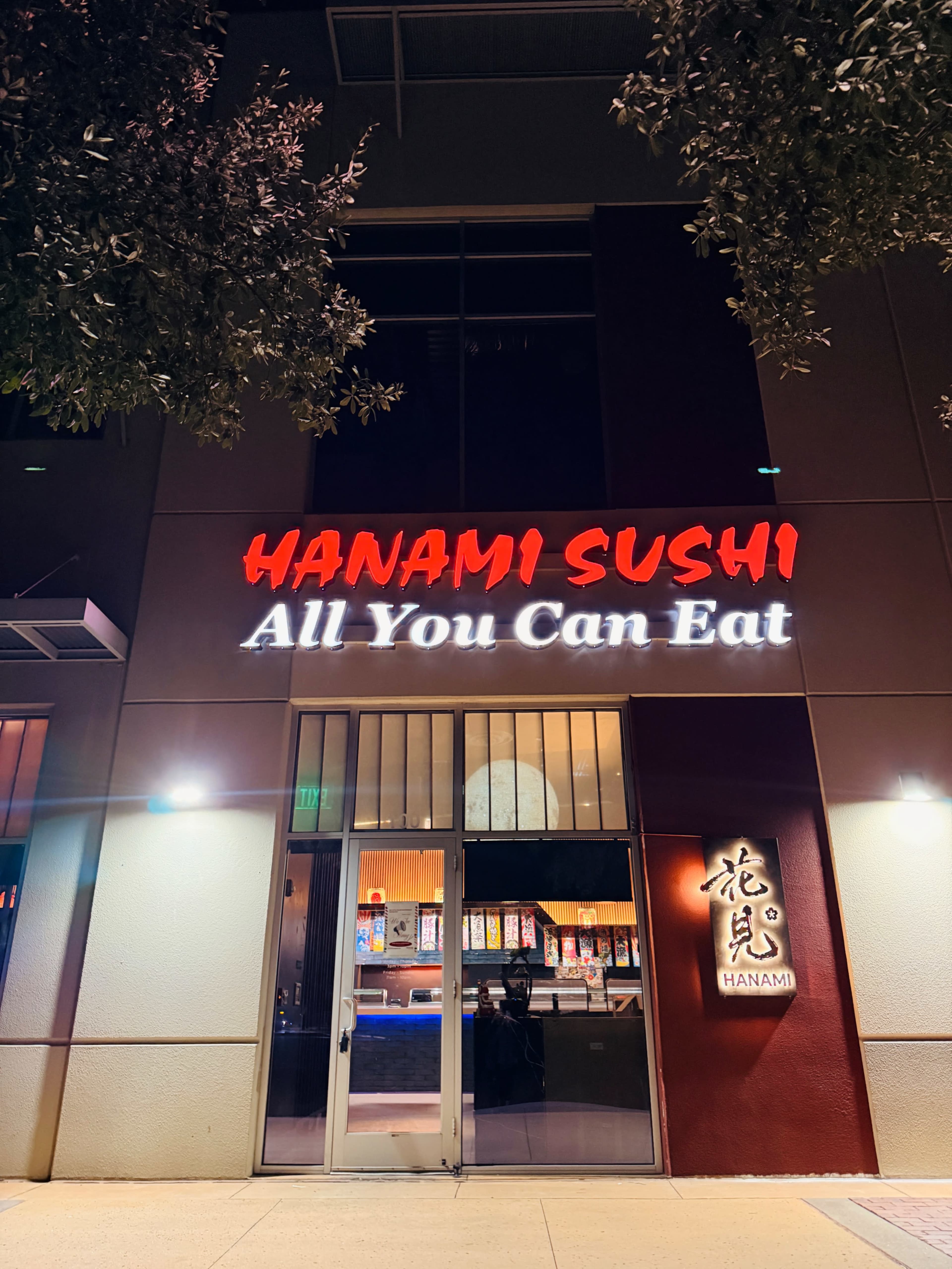 The storefront of Hanami Sushi, featuring a bright red sign that reads "HANAMI SUSHI" and "All You Can Eat," is illuminated at night.