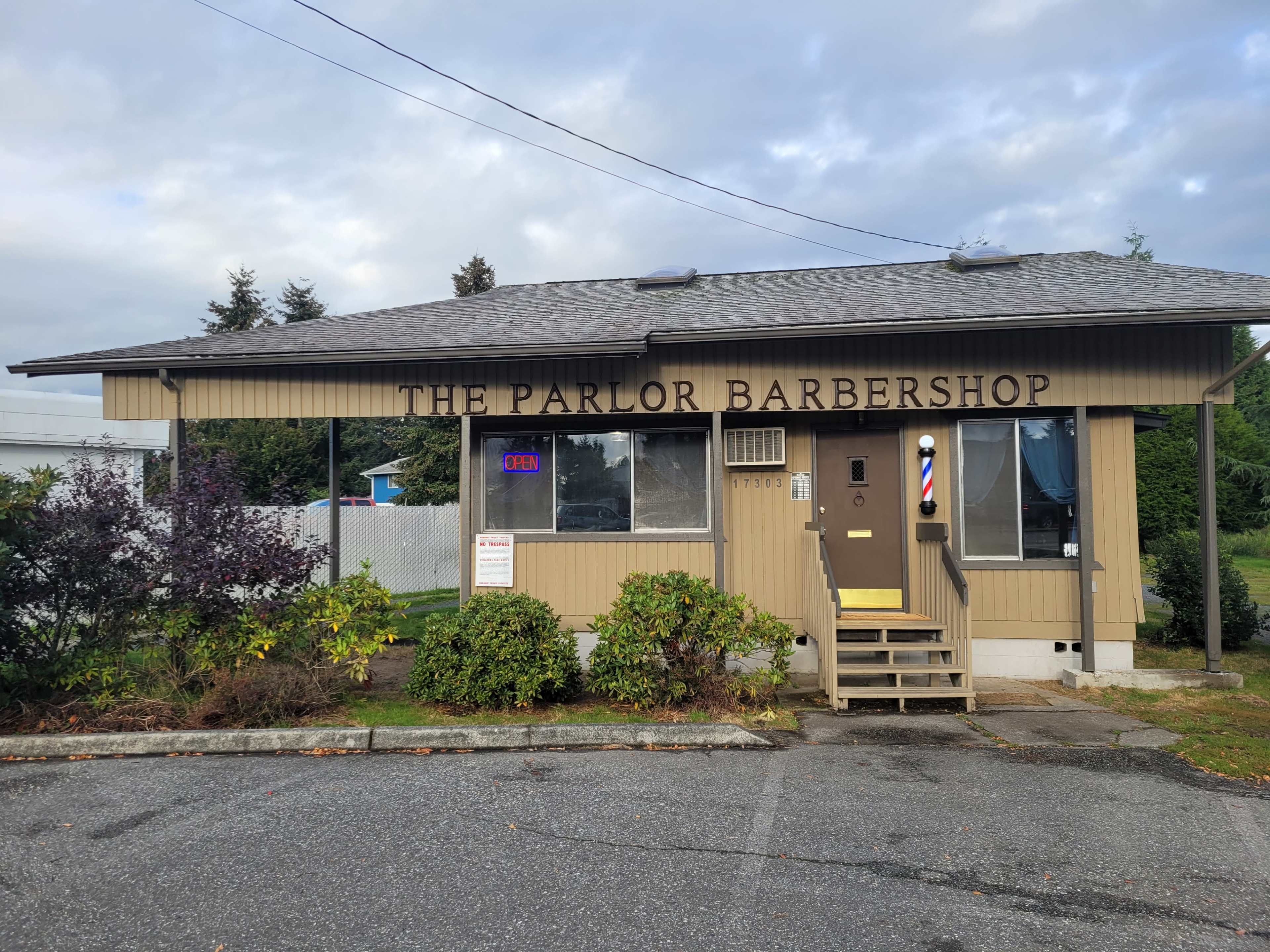 The image shows a one-story barbershop with a sign reading "The Parlor Barbershop" and a door featuring an open sign, located in a grassy area with some shrubs.