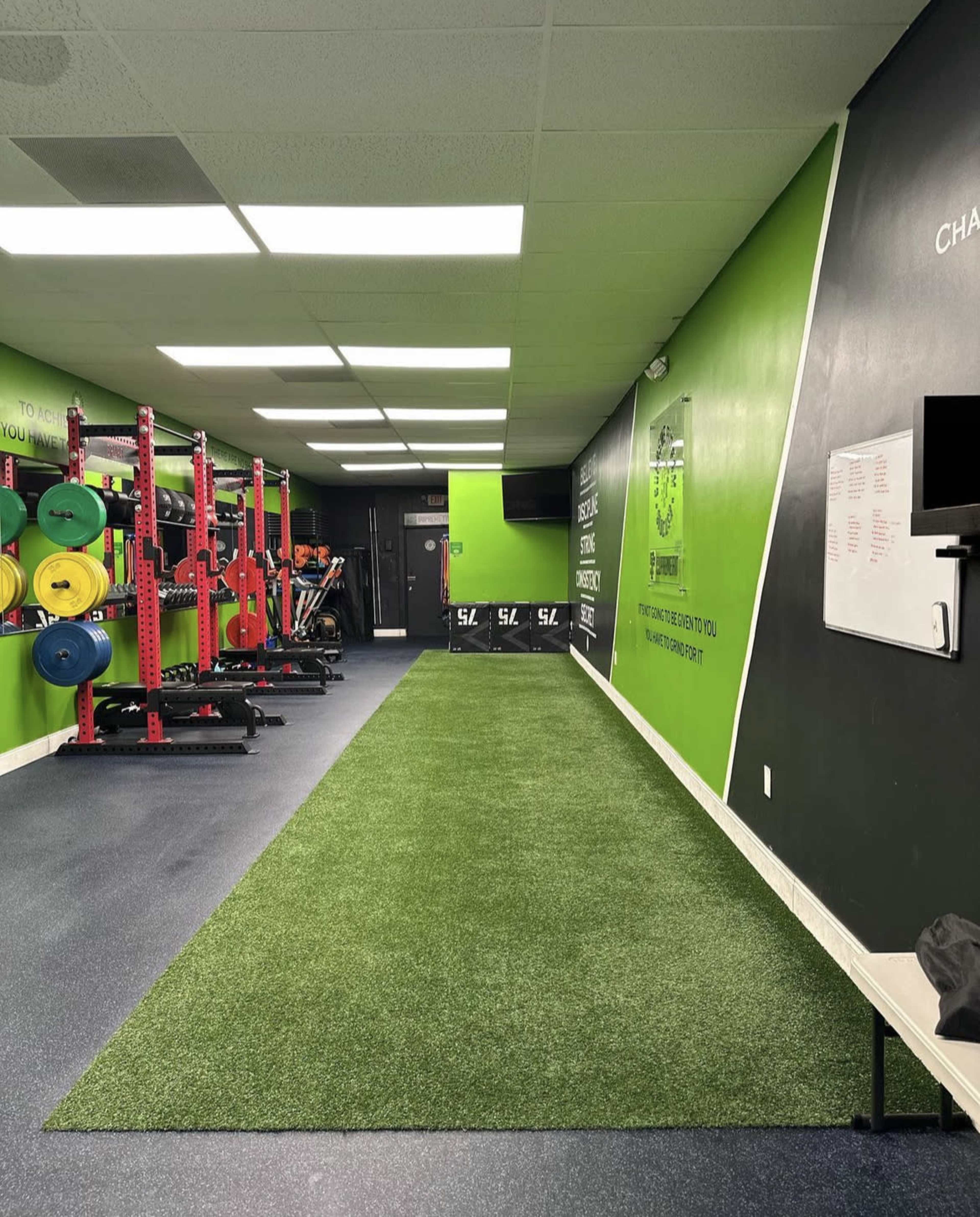 A fitness gym with a green turf area, weightlifting equipment, and a black wall with motivational quotes.