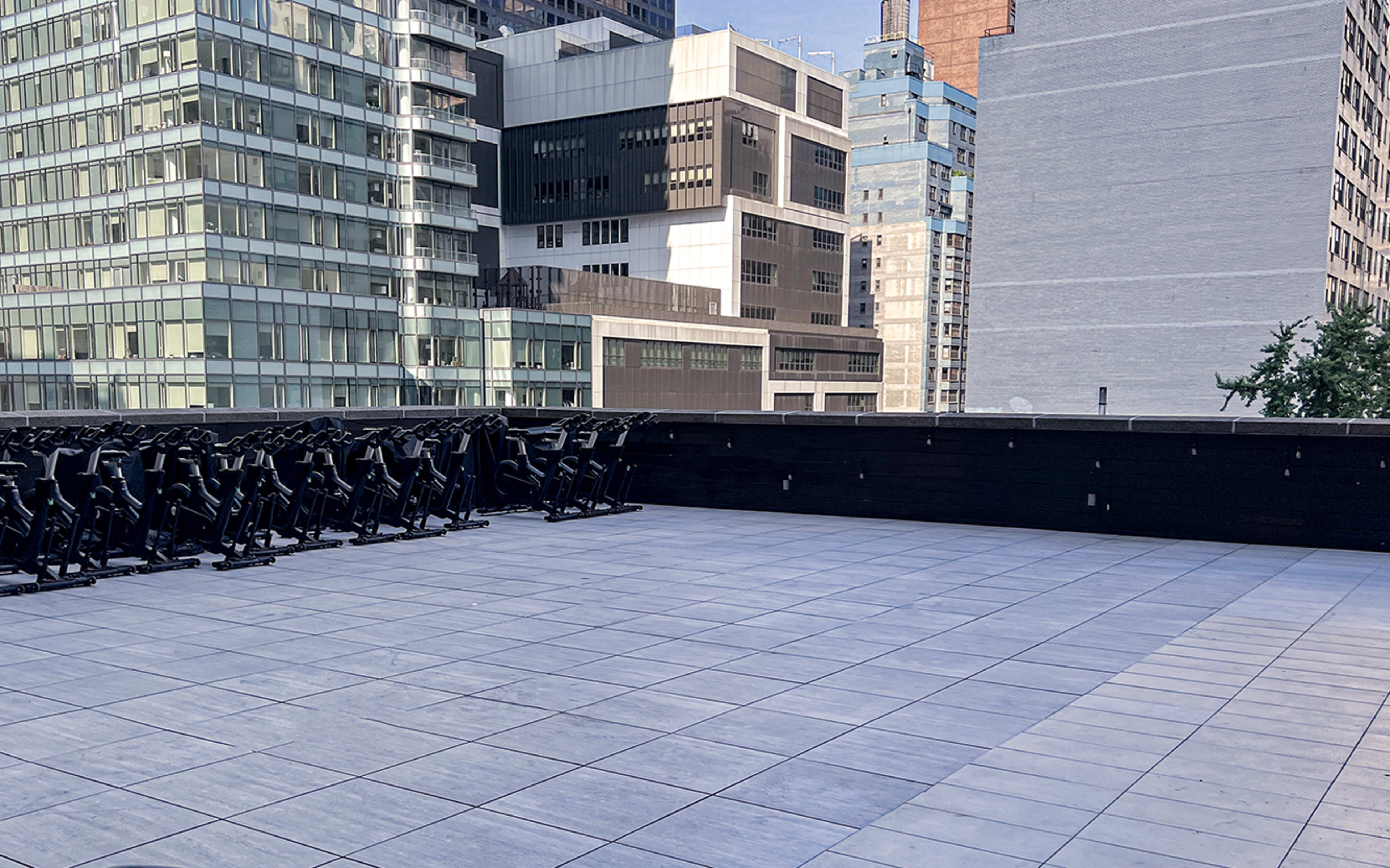 A rooftop terrace featuring a spacious tiled area with several black exercise bikes arranged against a backdrop of modern buildings.