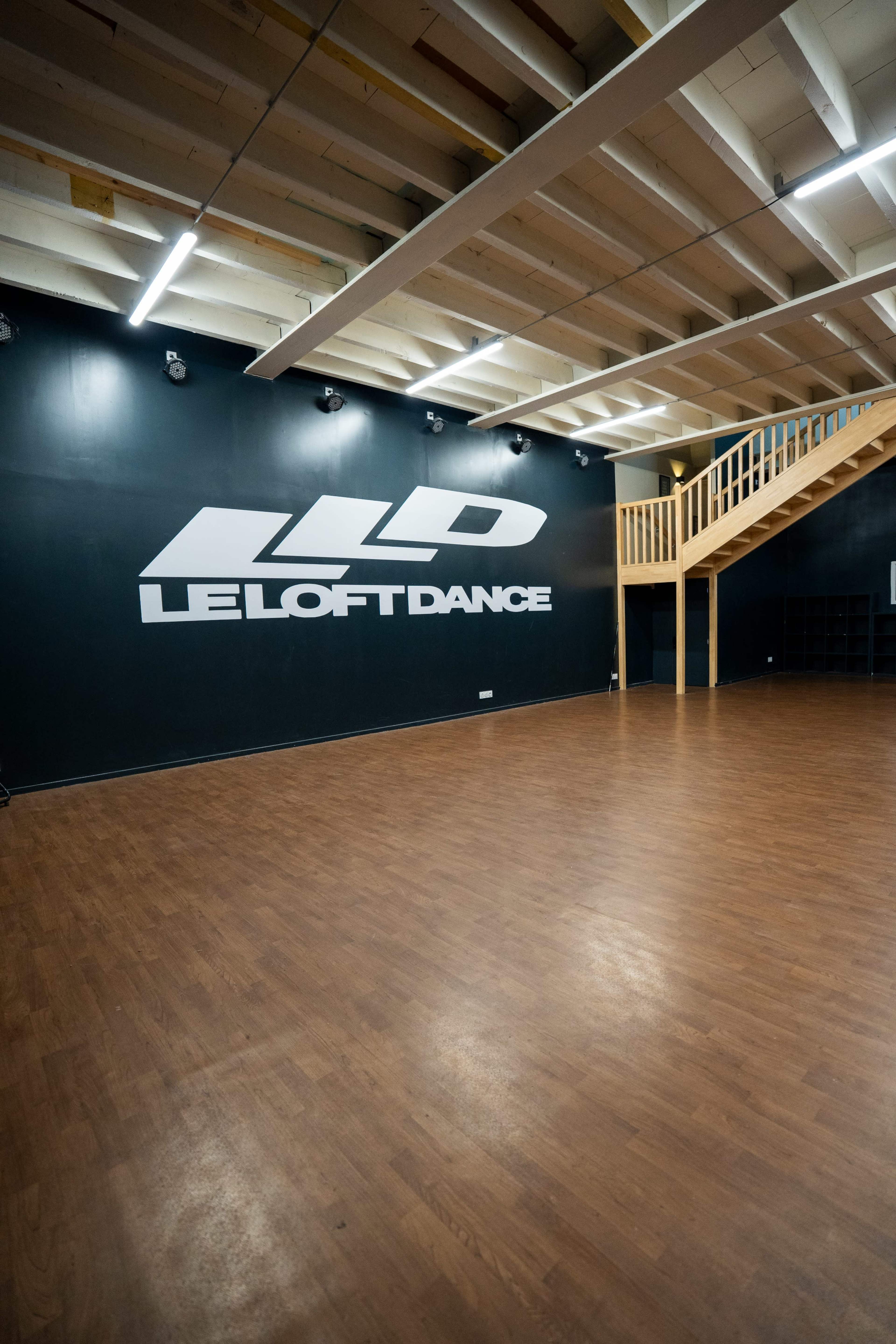 The image shows a spacious dance studio with a wooden floor, a black wall featuring the logo "LE LOFT DANCE," and an elevated wooden platform along one side.