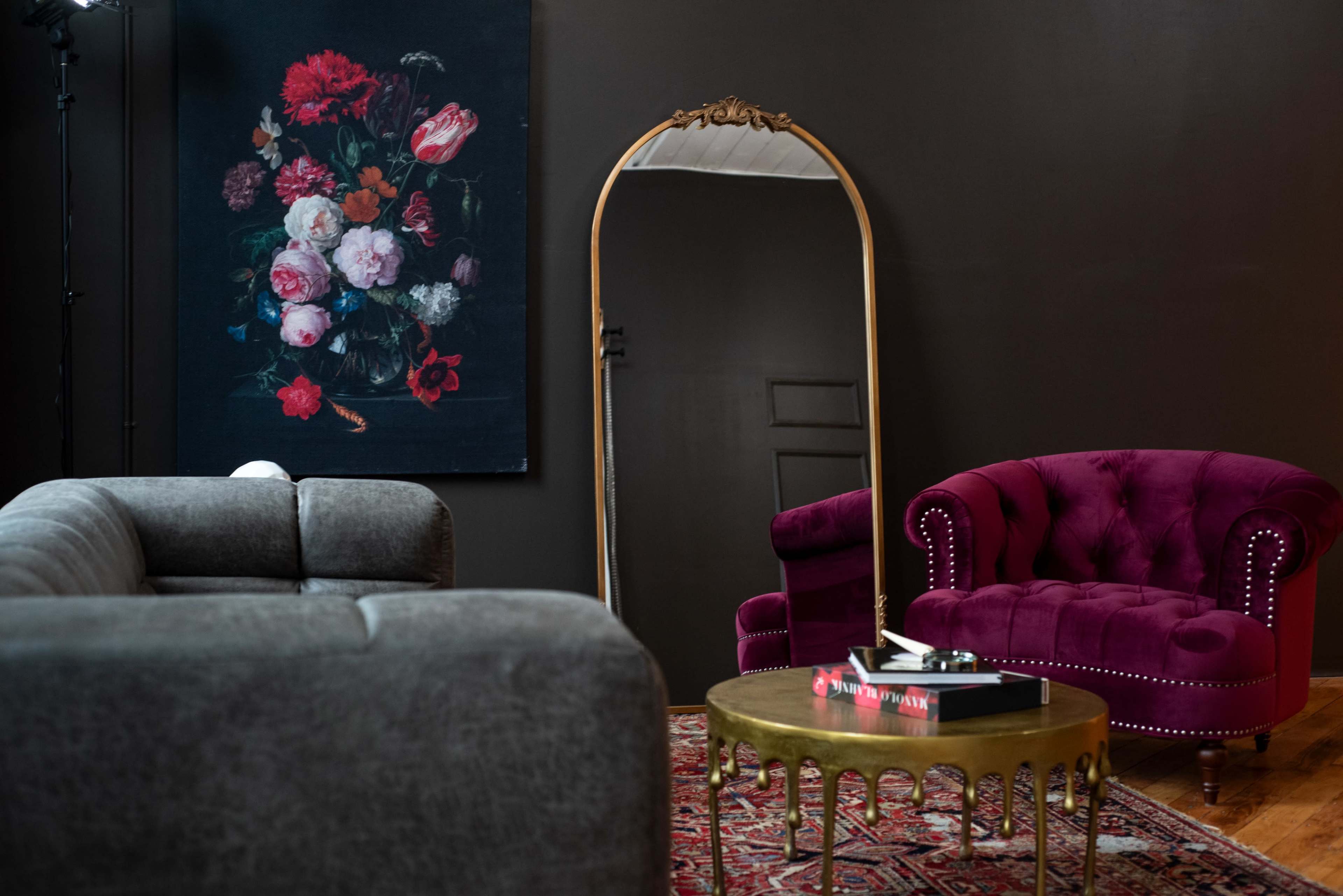 A modern living space features a grey sofa, a purple tufted armchair, a circular gold coffee table with books, and a large floral painting on the wall opposite an ornate mirror.