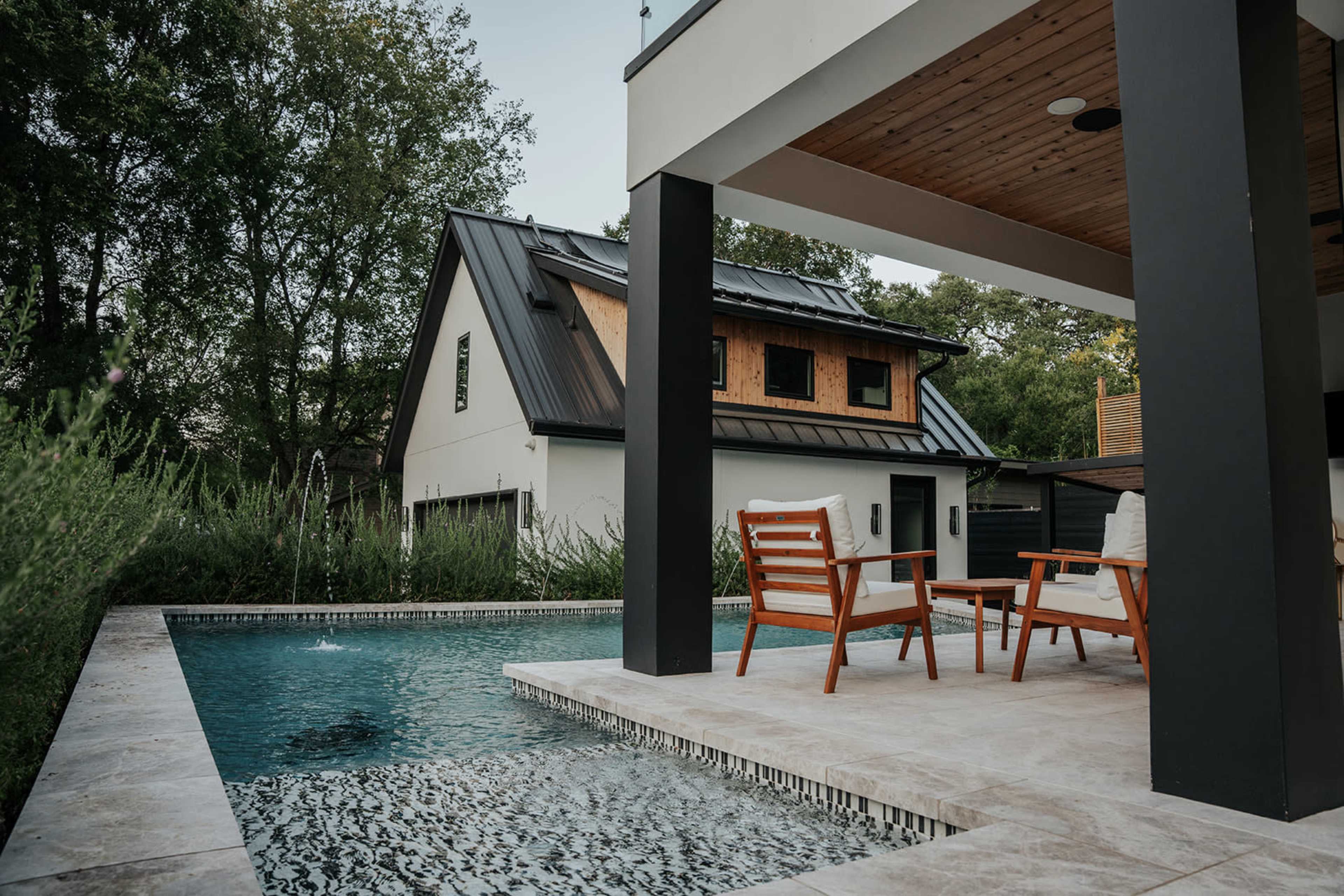 A modern home features a poolside seating area with wooden chairs and a backdrop of lush greenery.