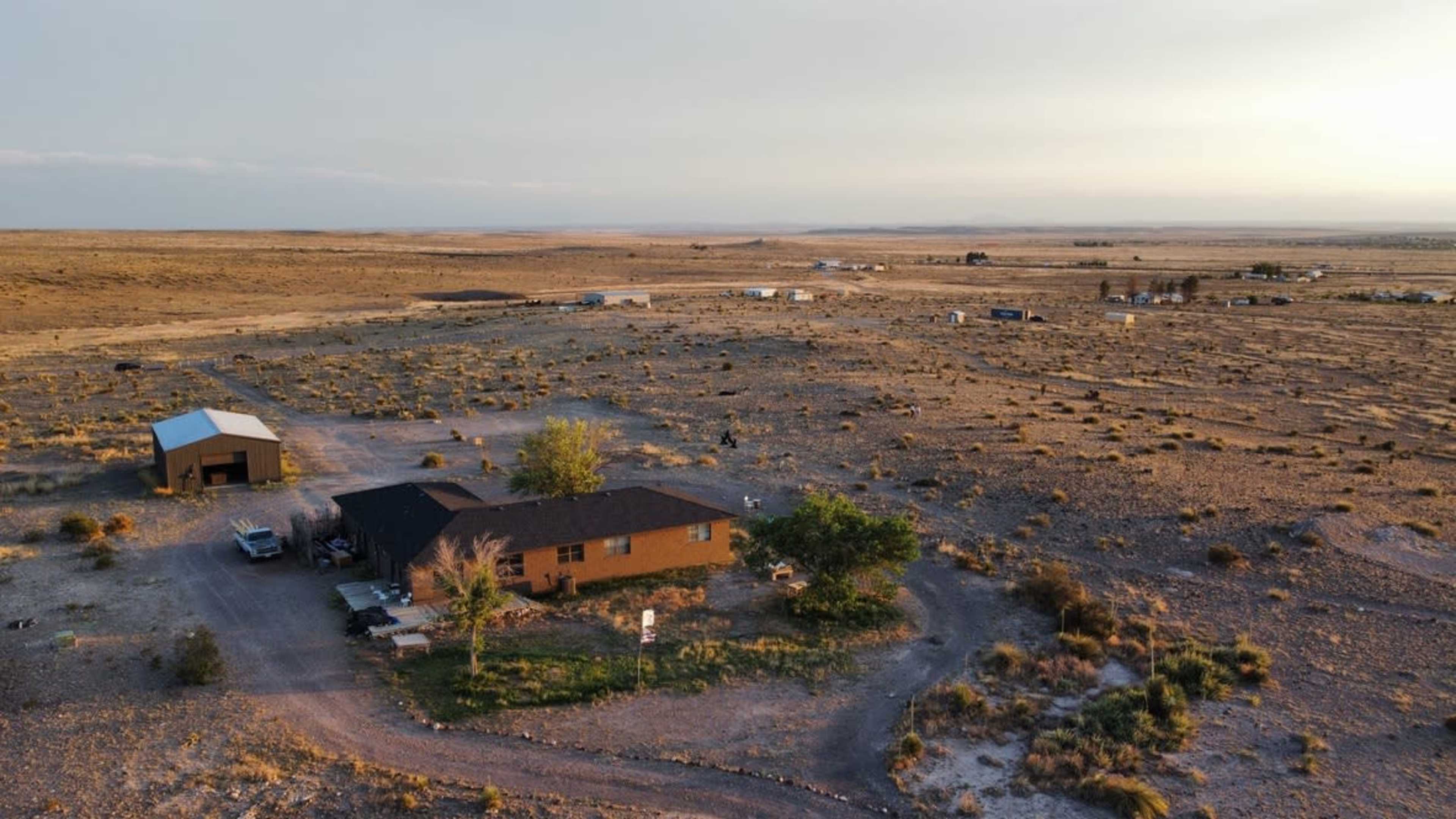 A single-story house with a dark roof sits amidst a sparse, arid landscape dotted with bushes and dirt roads.