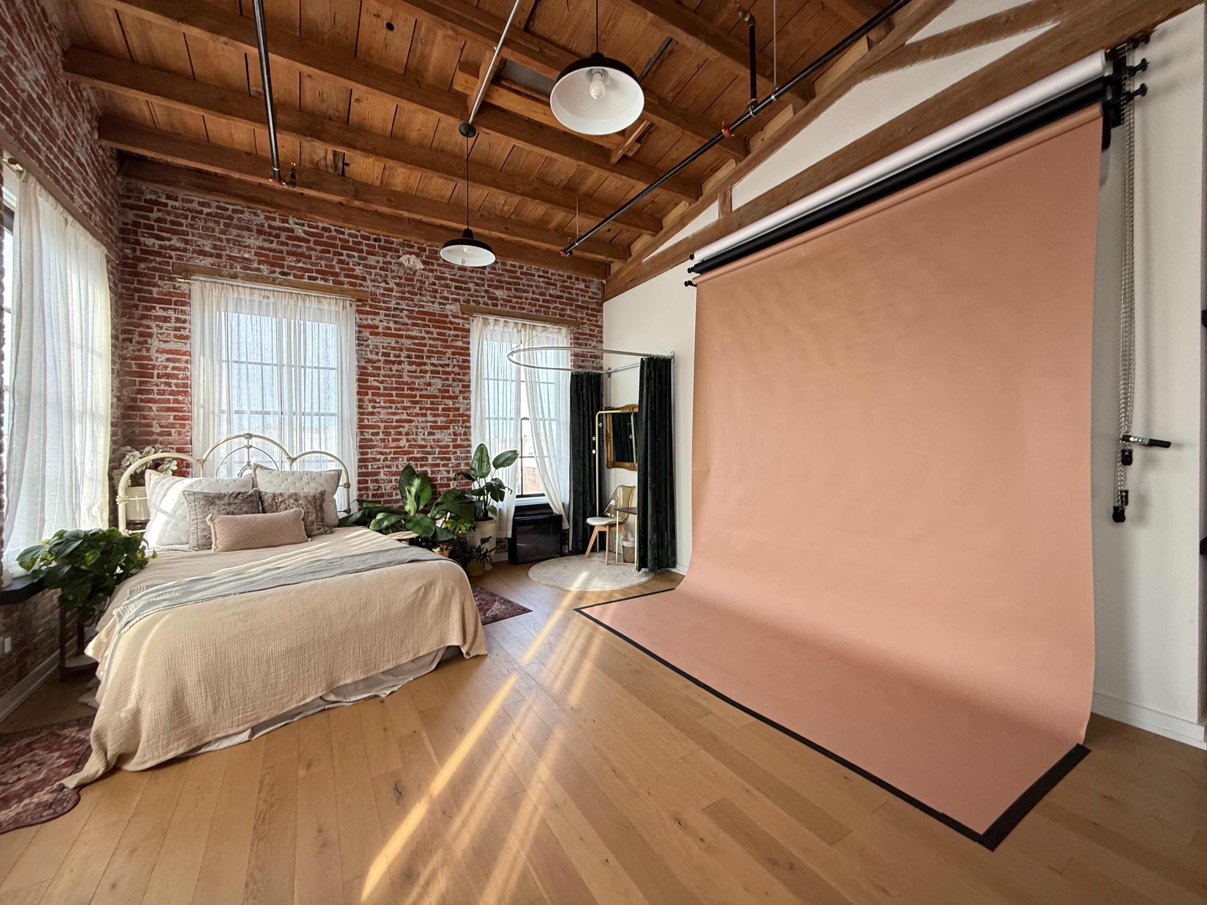 The image shows a cozy bedroom featuring a bed with neutral bedding, exposed brick walls, large windows, and a photography backdrop on one side.