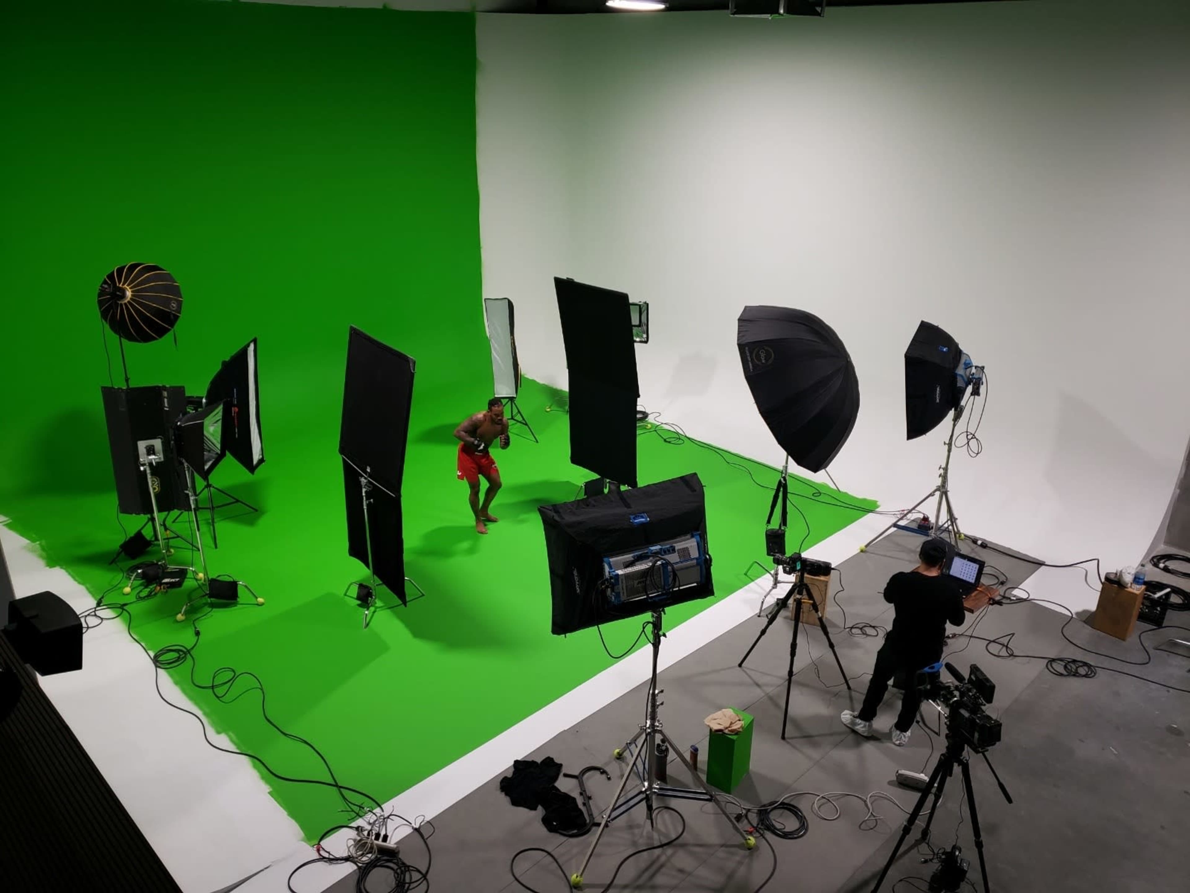 A person is posing on a green backdrop in a photo studio equipped with lighting and cameras.